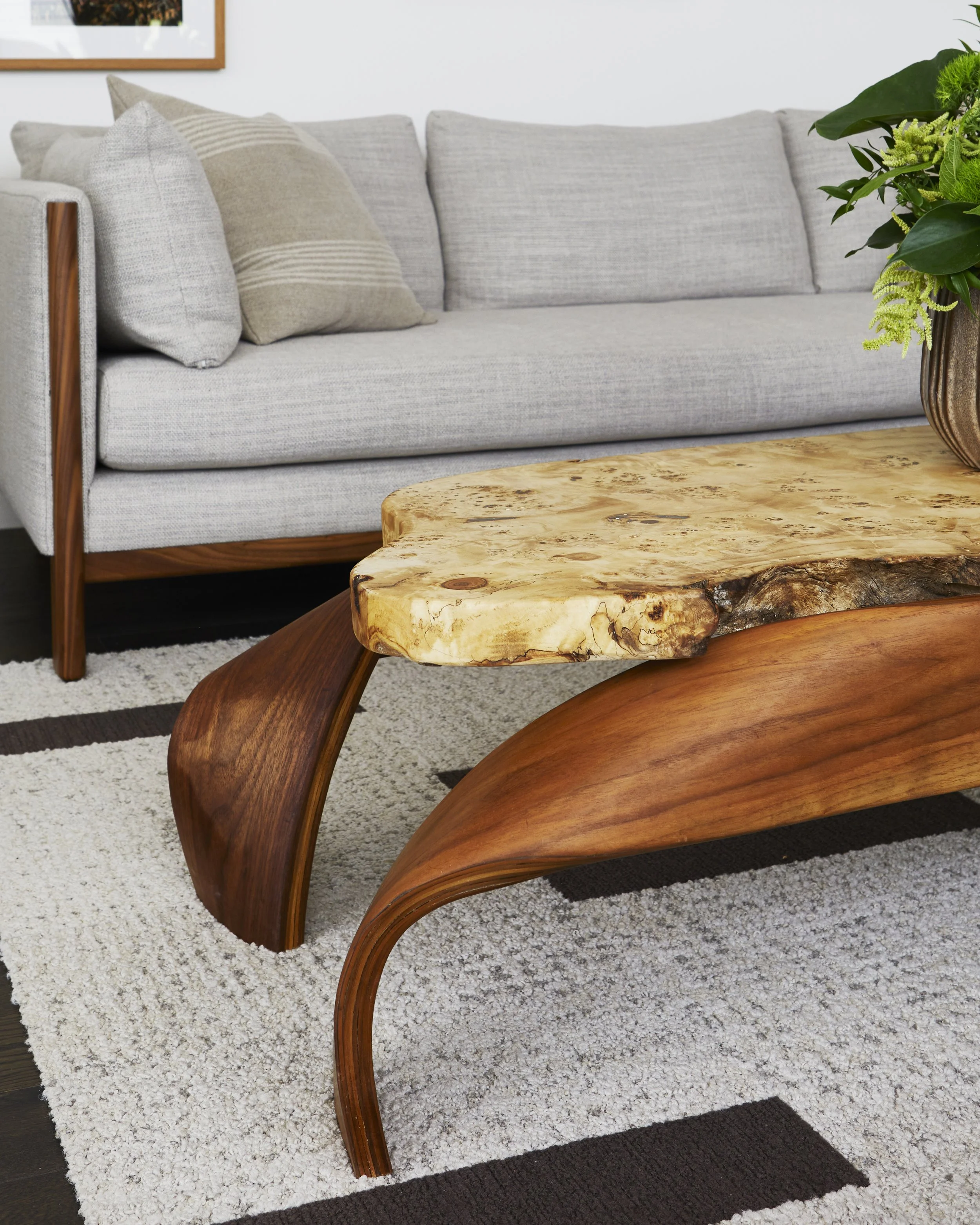 A living room with a light gray sofa, decorative pillows, a wooden coffee table with a live edge top, a patterned rug, and a potted plant.