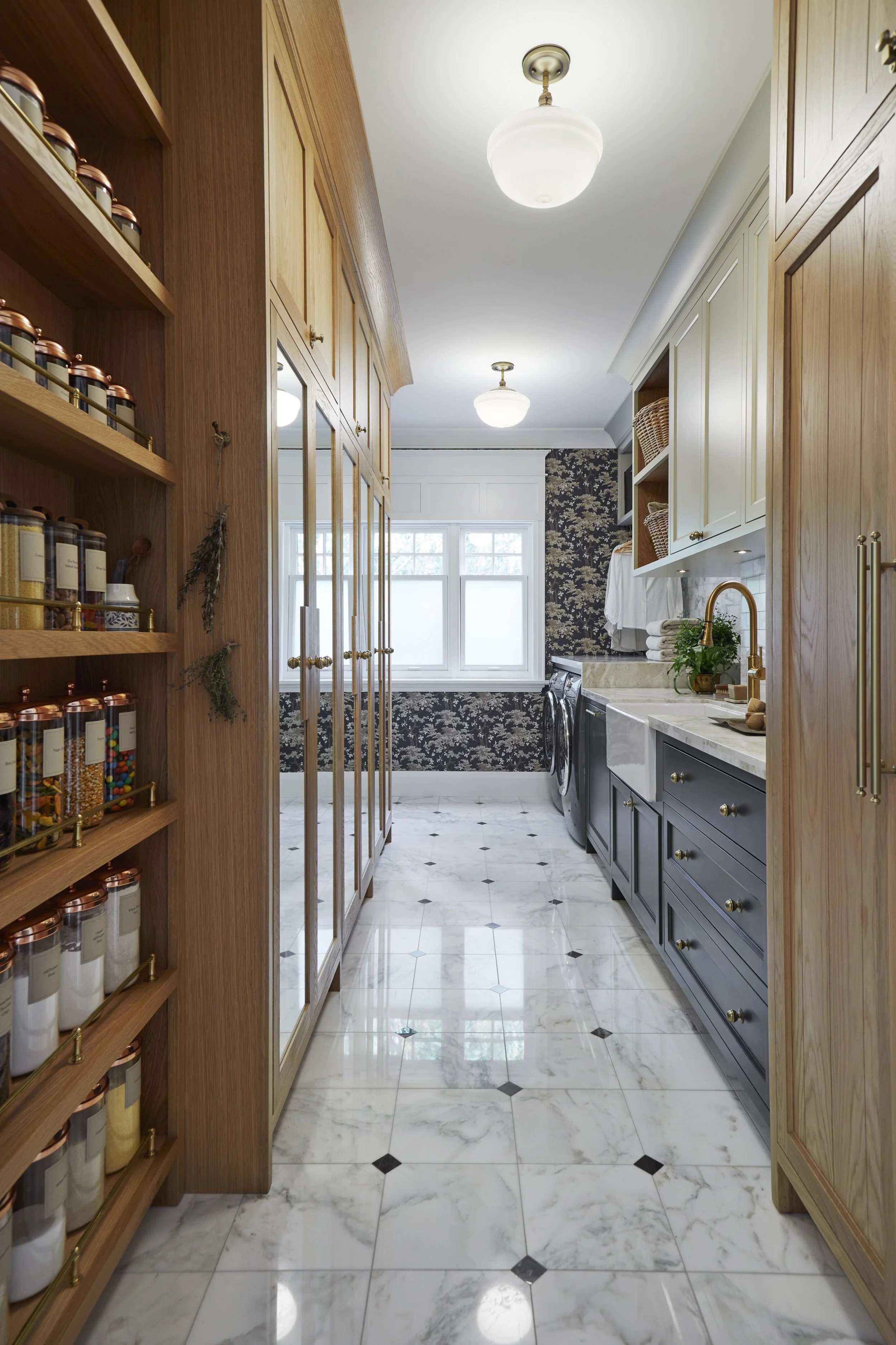 A spacious laundry room with white marble tile flooring, dark blue cabinets, beige upper cabinets, gold handles, a sink with a gold faucet, a window, and shelves with baskets. Wooden cabinetry and spice jars are on the left, and black and white patte