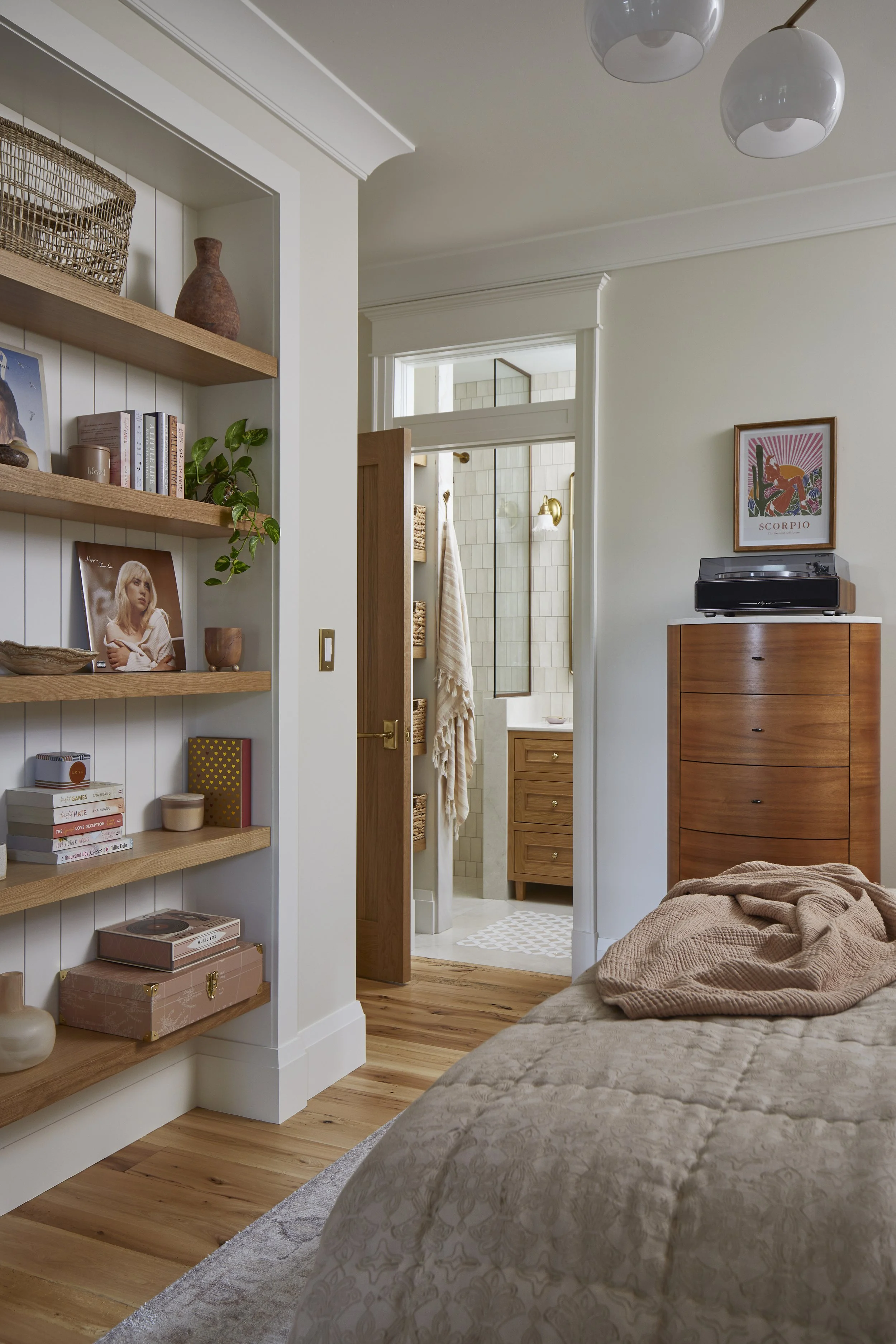 A cozy bedroom with a built-in wooden bookshelf, a dresser with a music player, and a partially opened door leading to a bathroom with a vanity and towels.