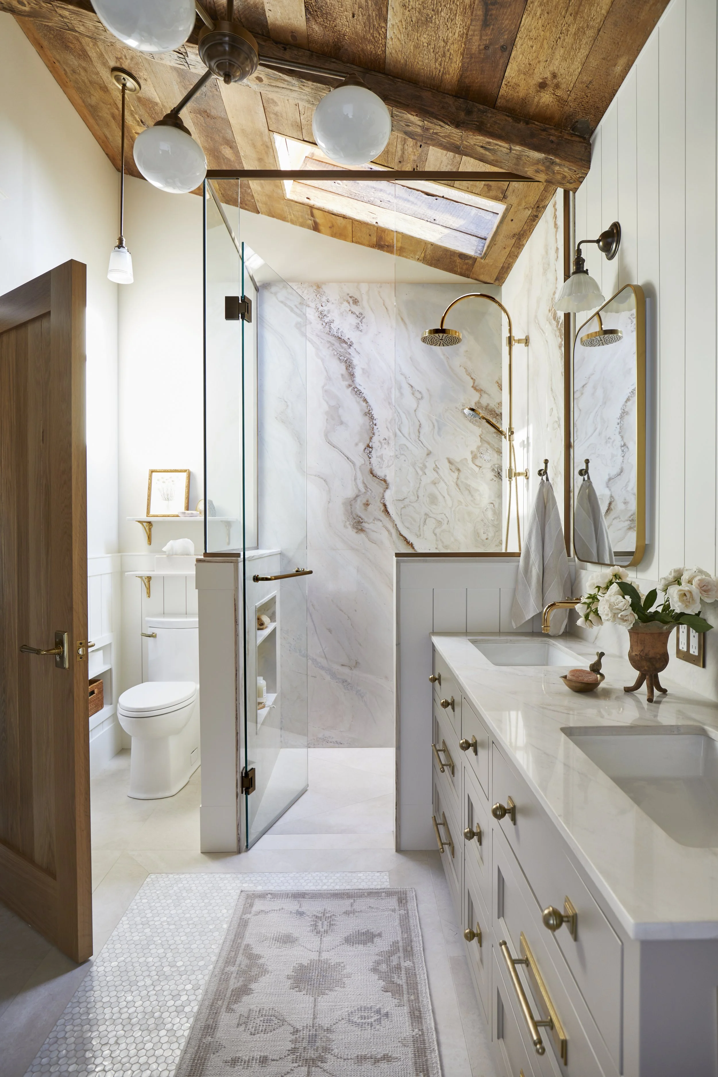 A modern bathroom with double sinks, a large mirror, a shower with a gold showerhead, a skylight, and a toilet, featuring white cabinetry, gold fixtures, and decorative accents.