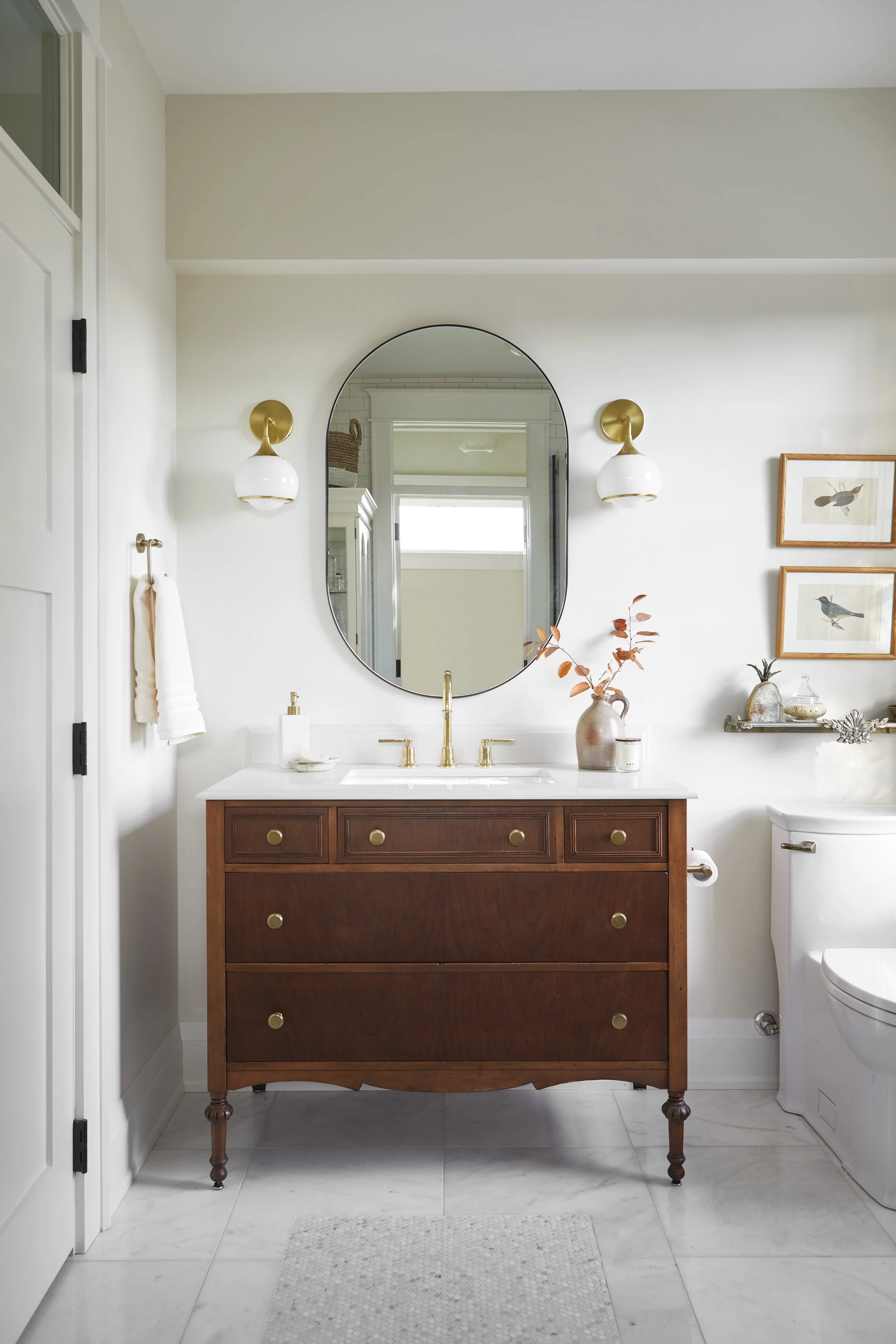 A bathroom with a wooden vanity, a large oval mirror, wall-mounted lights, a vase with flowers, framed bird pictures, and a toilet.