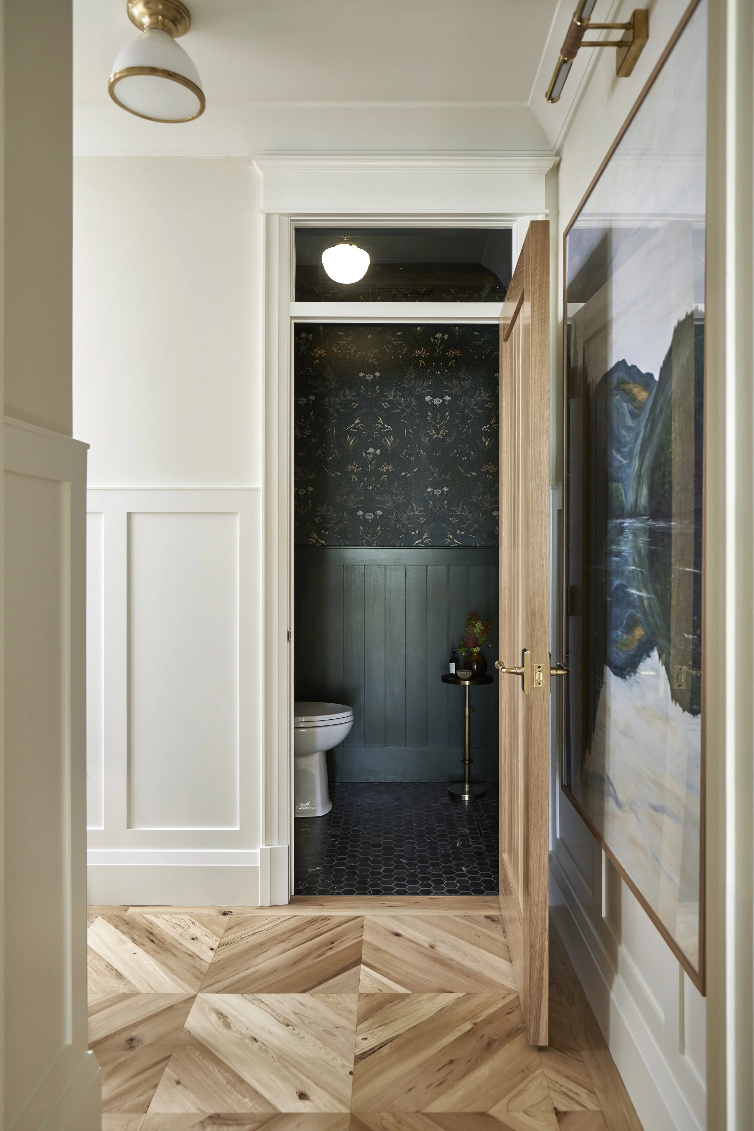 View of a hallway leading to a small bathroom with dark wallpaper and green paneling, featuring a white toilet and a small side table with decorations.
