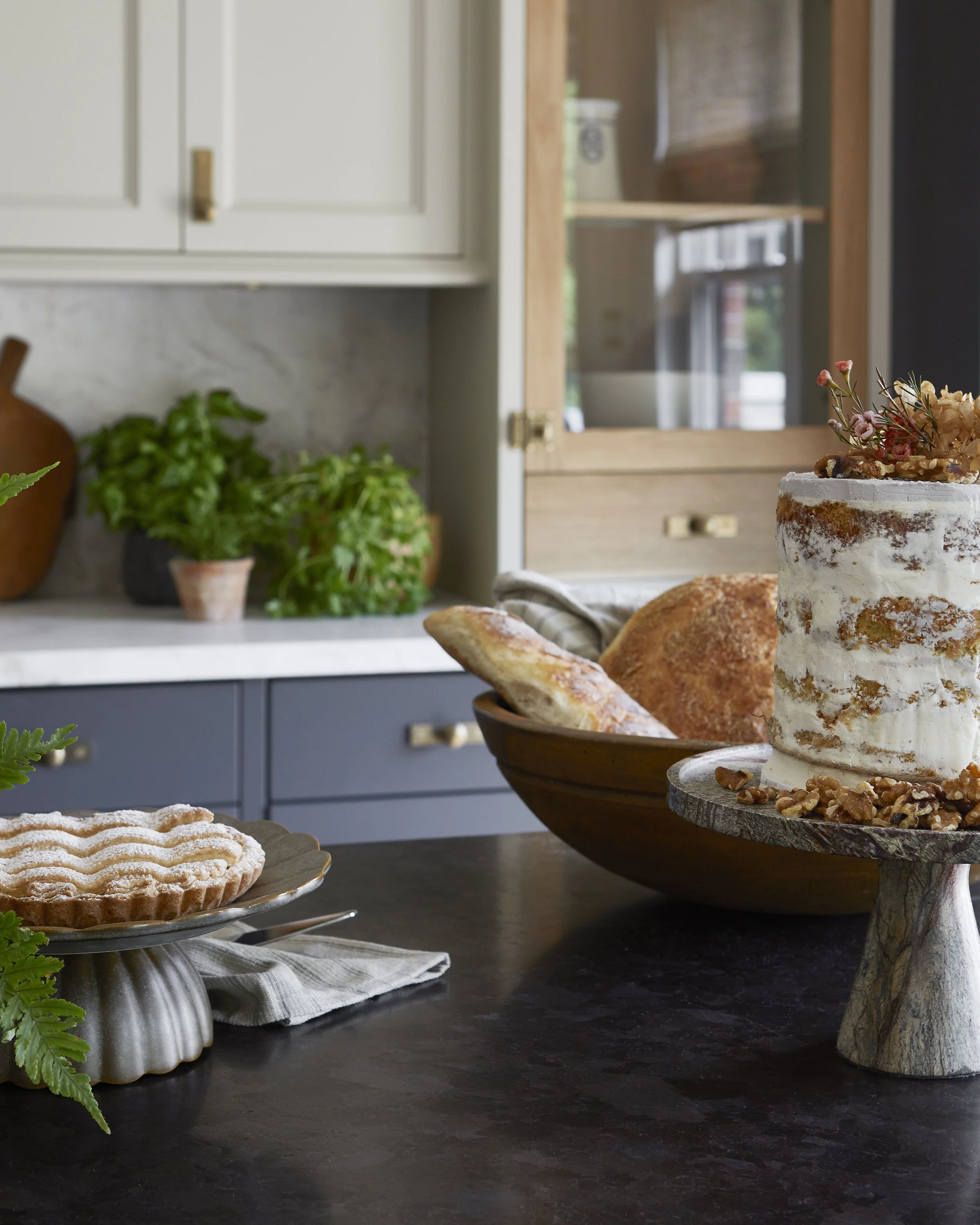A bakery setup with a tall white layered cake decorated with flowers on a stone cake stand, a pie on a silver cake stand, and a bowl of bread on a wooden bowl in a kitchen with green plants and wooden cabinets.
