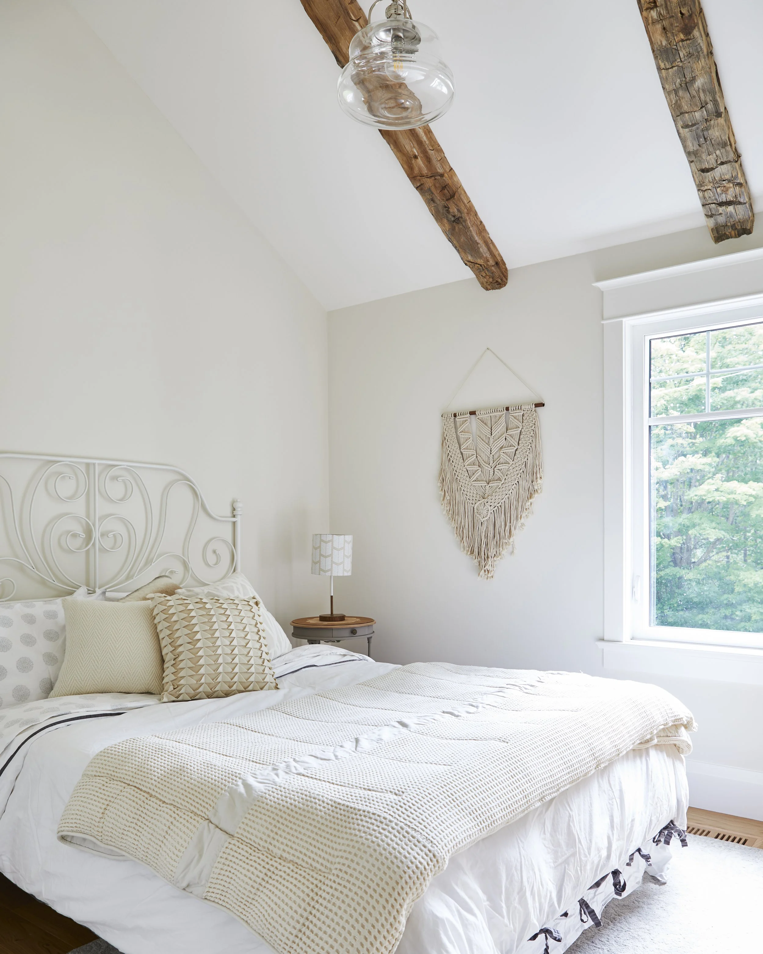 Bedroom with white walls, rustic wooden beams on the ceiling, a window with greenery outside, a cream-colored bed with pillows, a nightstand with a lamp, and a wall hanging decor.