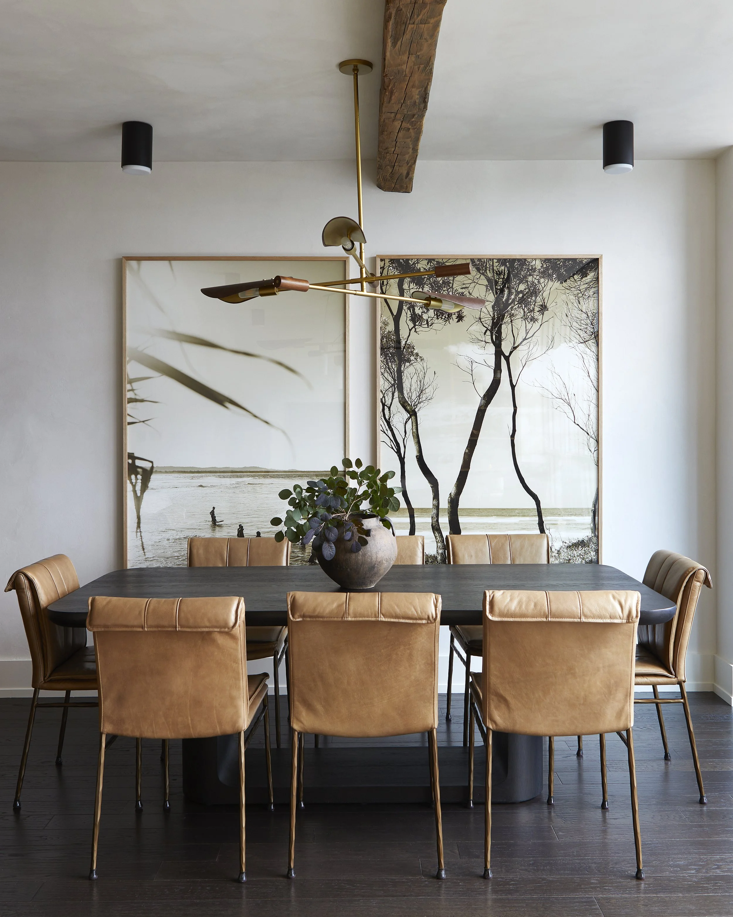 Modern dining room with a dark wooden table, tan leather chairs, two large framed nature-themed artworks on the wall, a gold and wood multi-armed pendant light, a potted plant centerpiece, dark hardwood floor, and white walls.