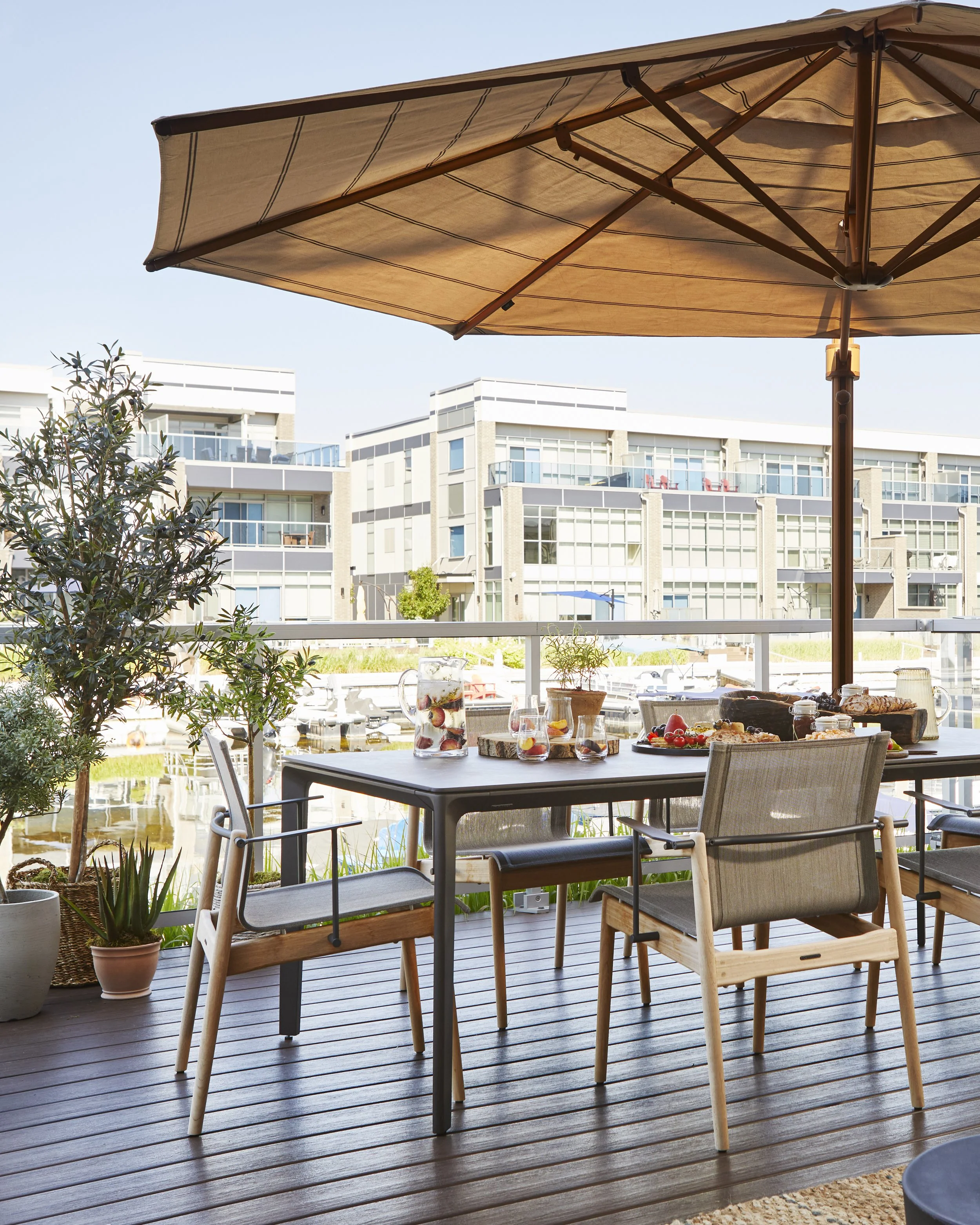 Outdoor patio with wooden flooring, a table set with drinks and snacks, chairs, potted plants, a large beige umbrella, with modern residential buildings in the background.
