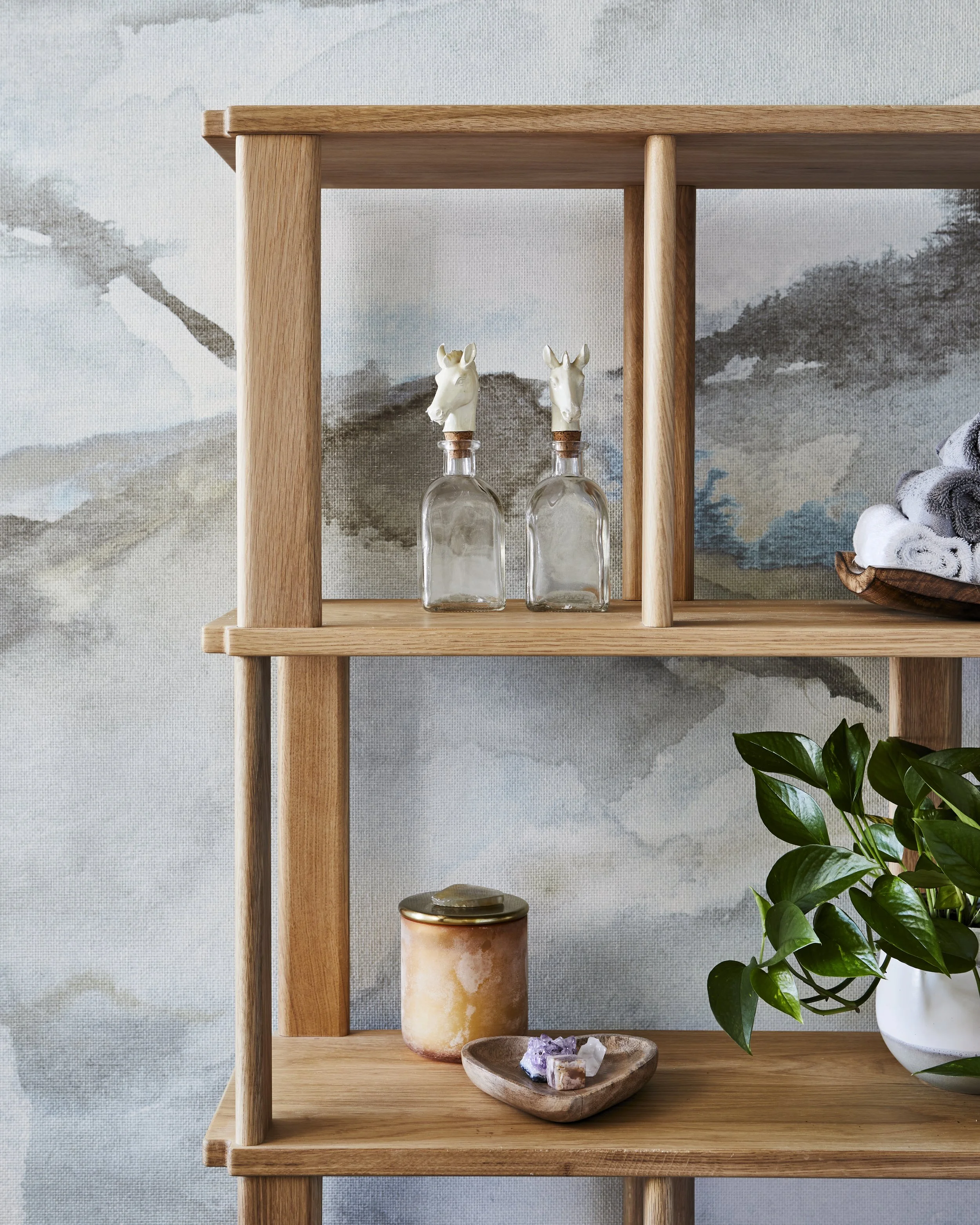Wooden shelf with decorative bottles topped with horse head stoppers, candles, rocks, and a potted plant against a watercolor-style landscape wall.