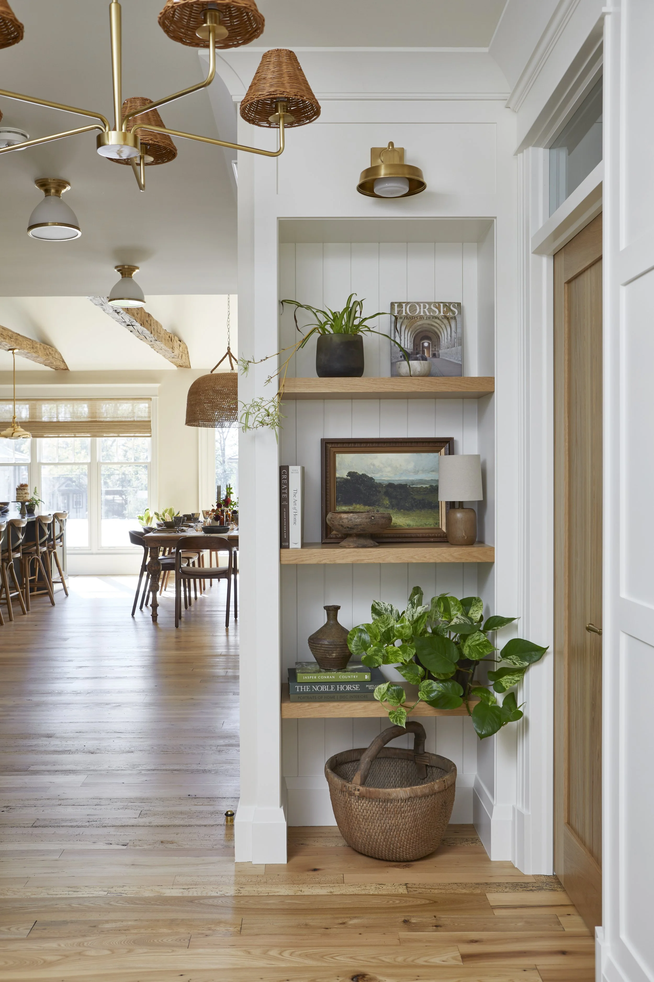 Built-in wall shelves with potted plants, books, and art in a room with hardwood floors and white walls, with a dining area visible in the background.
