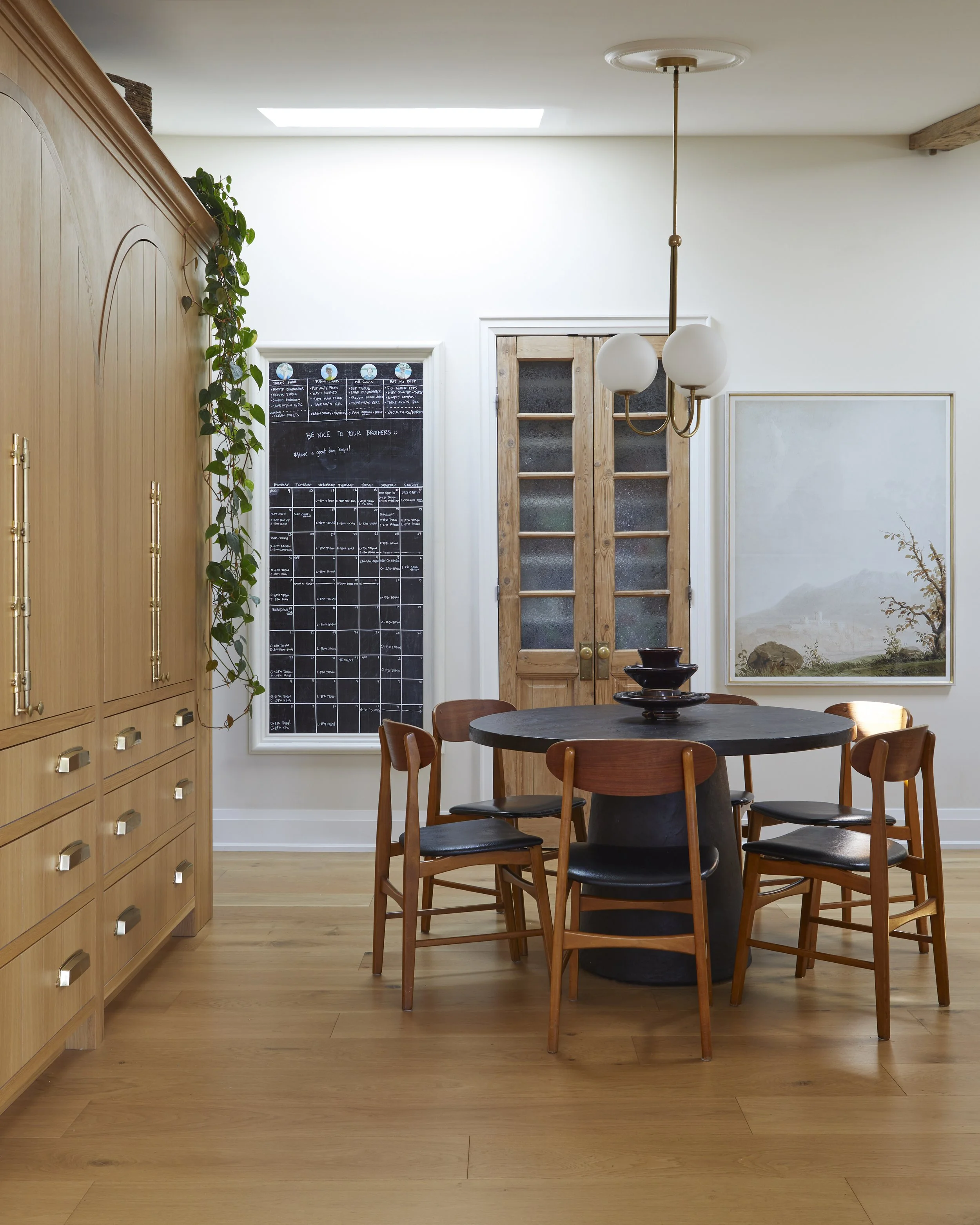 A dining room with wooden furniture, including a round table with six chairs, a large wooden cabinet on the left, a doorway with glass panels, a black chalkboard wall, a skylight, a painting on the wall, and a hanging light fixture.
