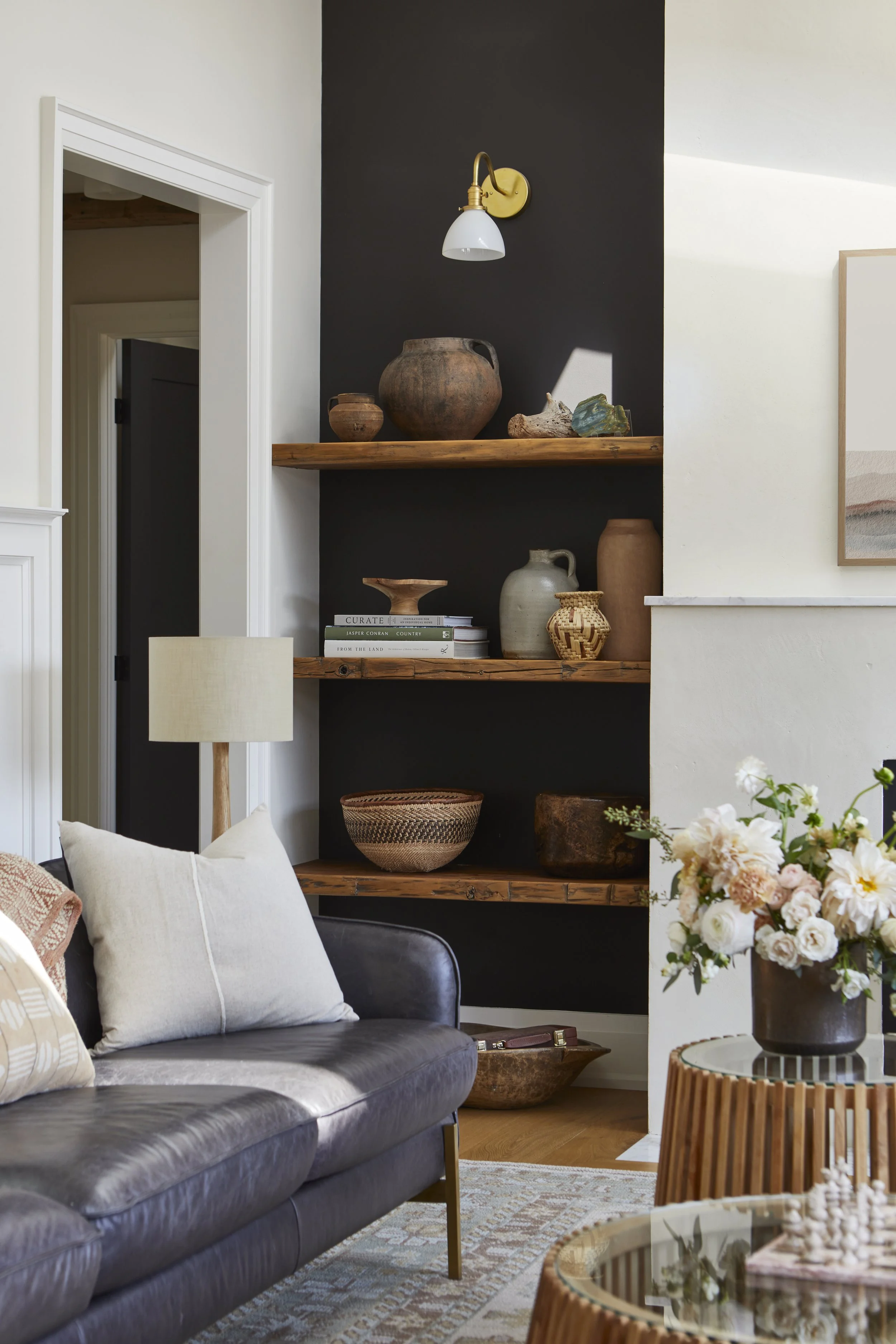 Living room with a black leather sofa, wooden shelves with pottery and books, a table with a flower arrangement, and a wall-mounted light fixture.