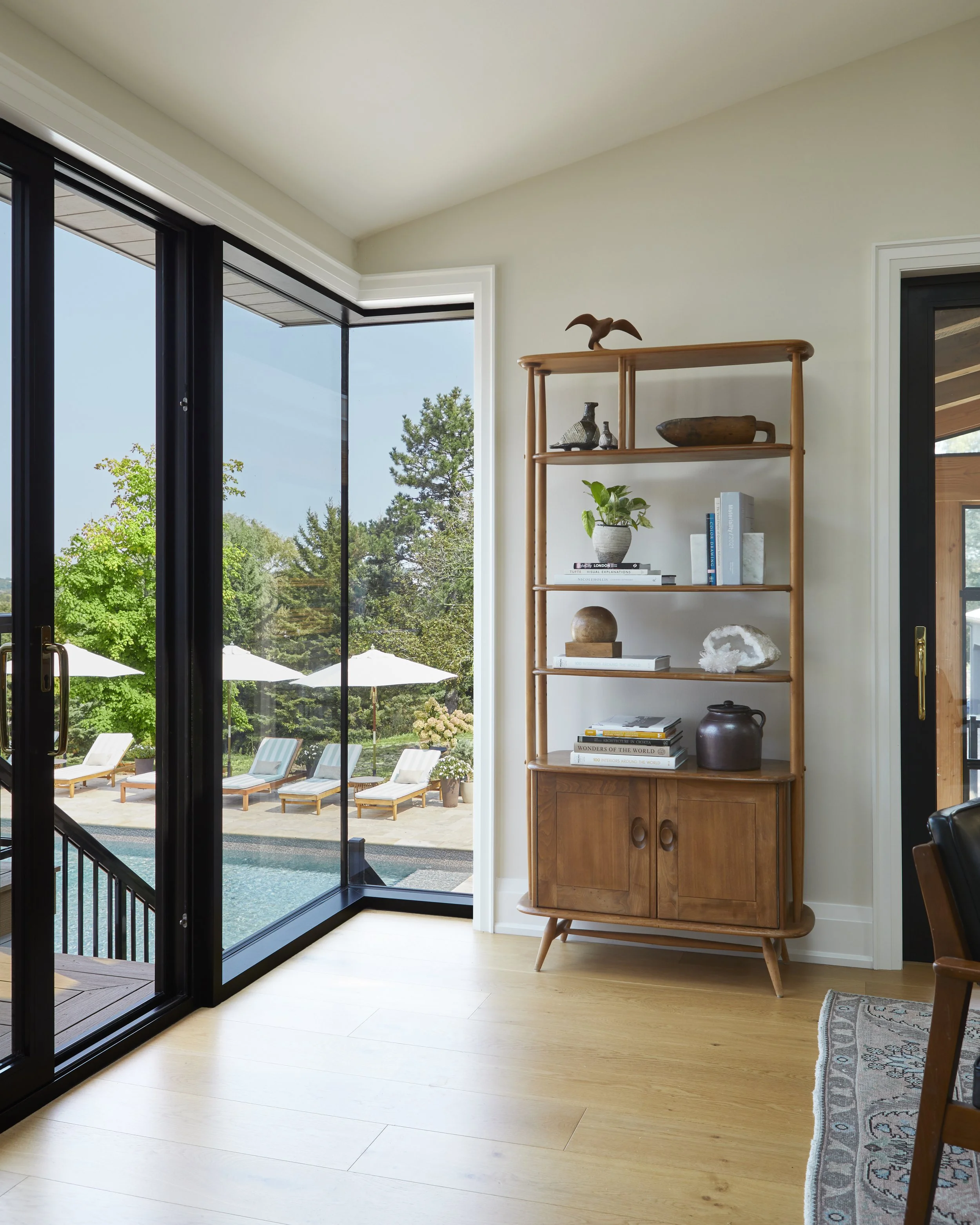 Interior view of a room with large glass doors leading to a patio with pool and lounge chairs, a wooden shelf with decorative items and plants, and a black sliding door.