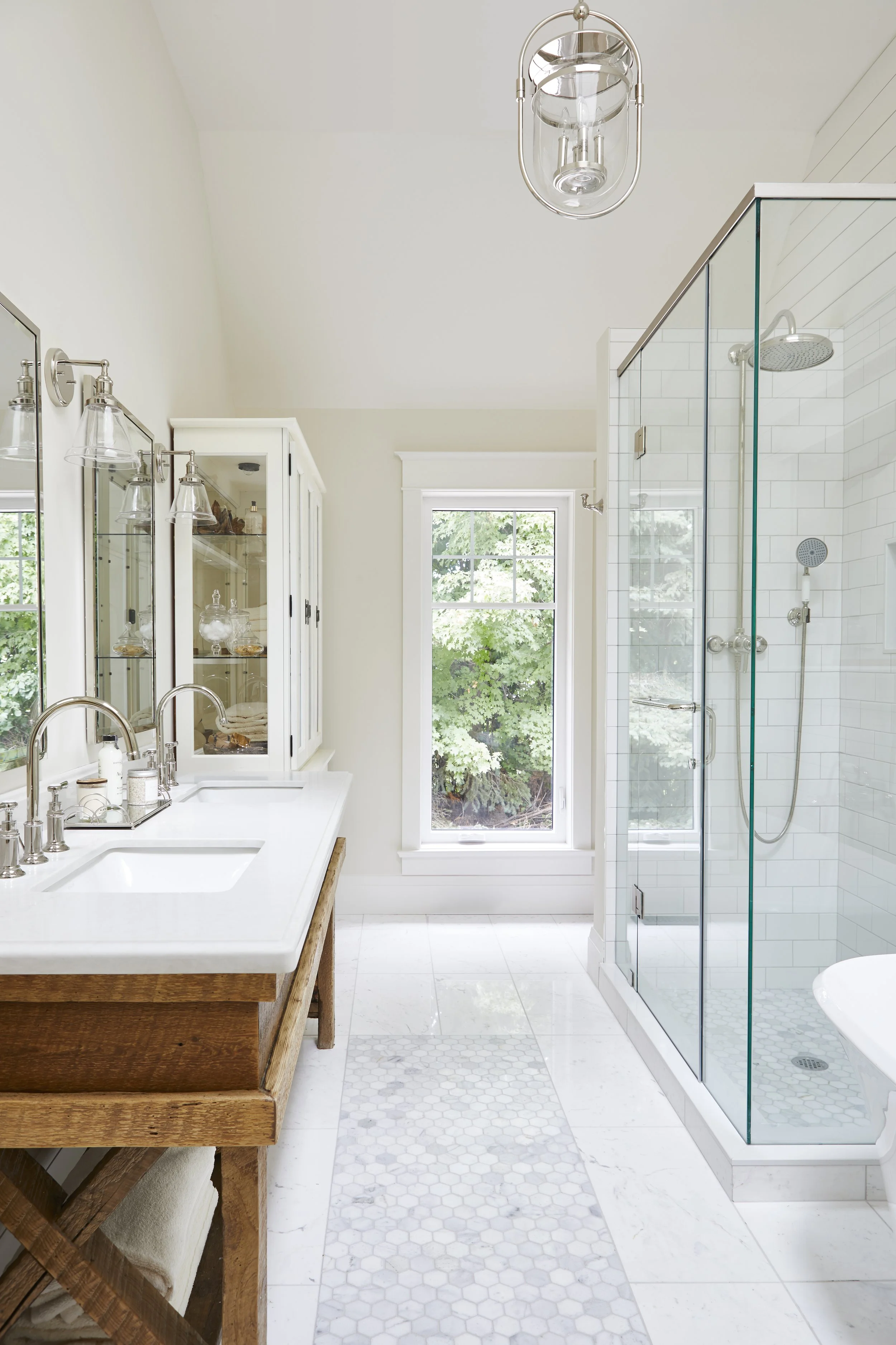 Bright bathroom with a wooden vanity, twin sinks, glass-fronted cabinet, window with greenery outside, and a glass-walled shower with white tile inside.