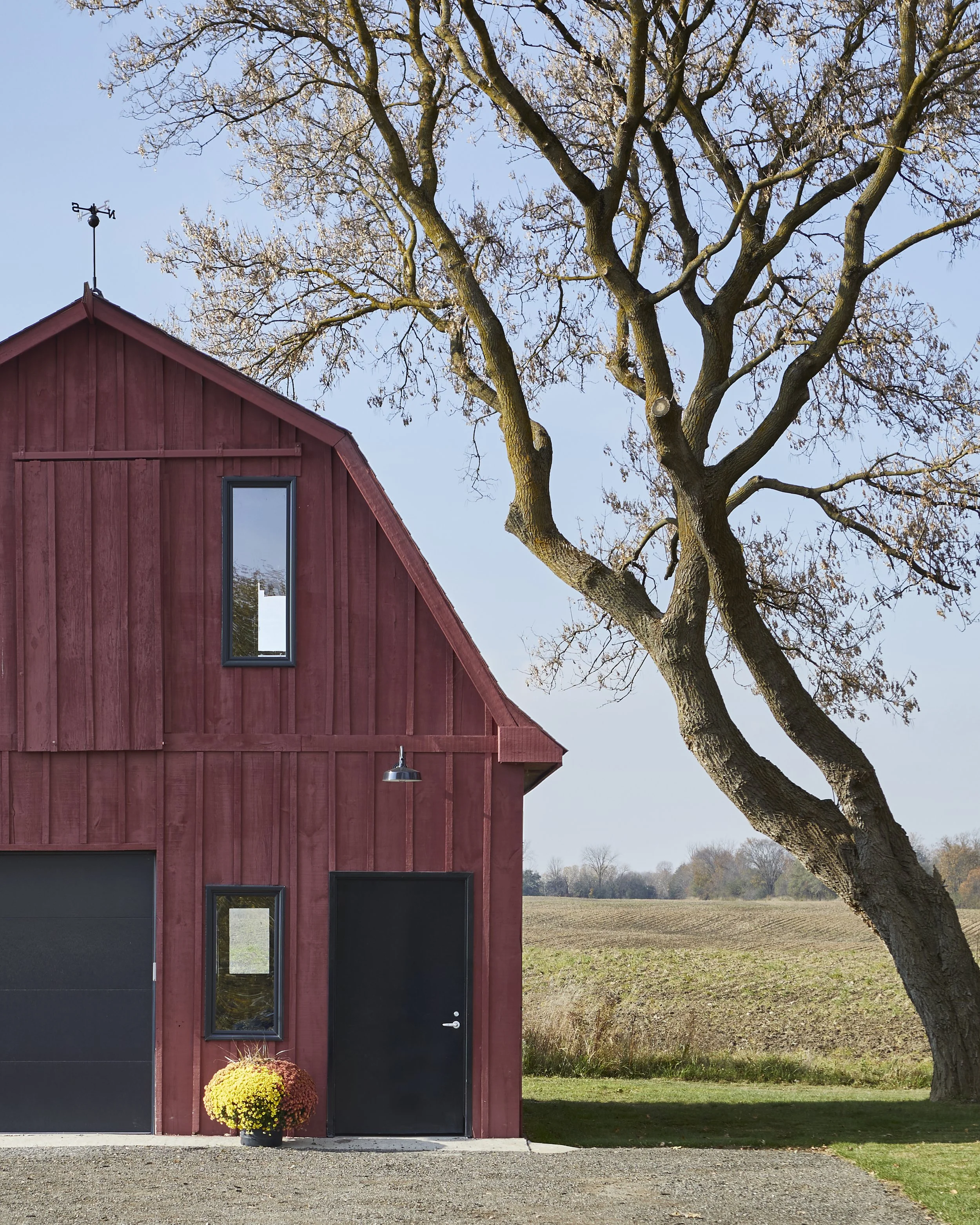 Red barn with a black door and windows, next to a leafless tree, in a rural landscape under a clear blue sky.