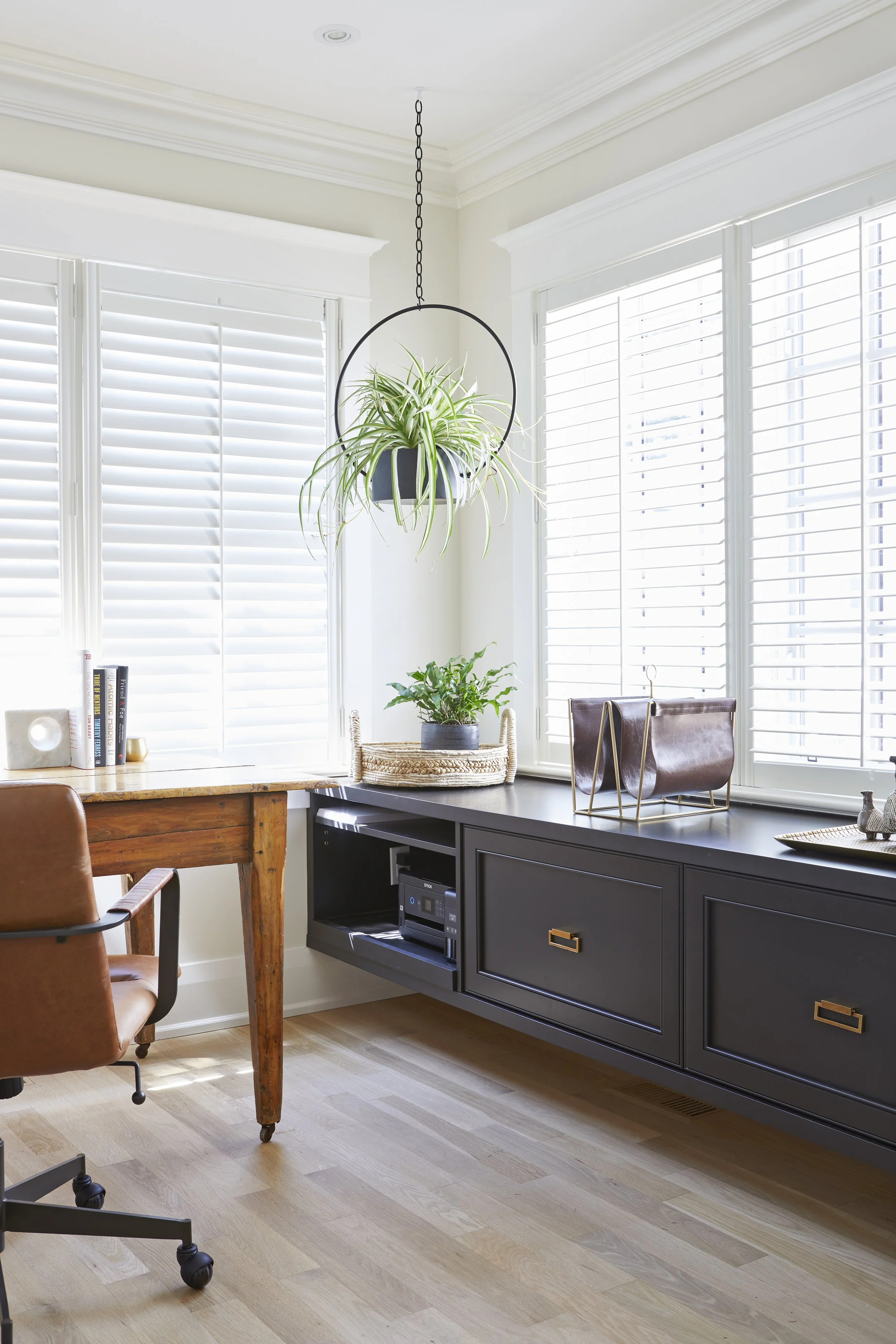 Bright home office corner with large windows, black sideboard, hanging potted plant, and desk with leather chair.