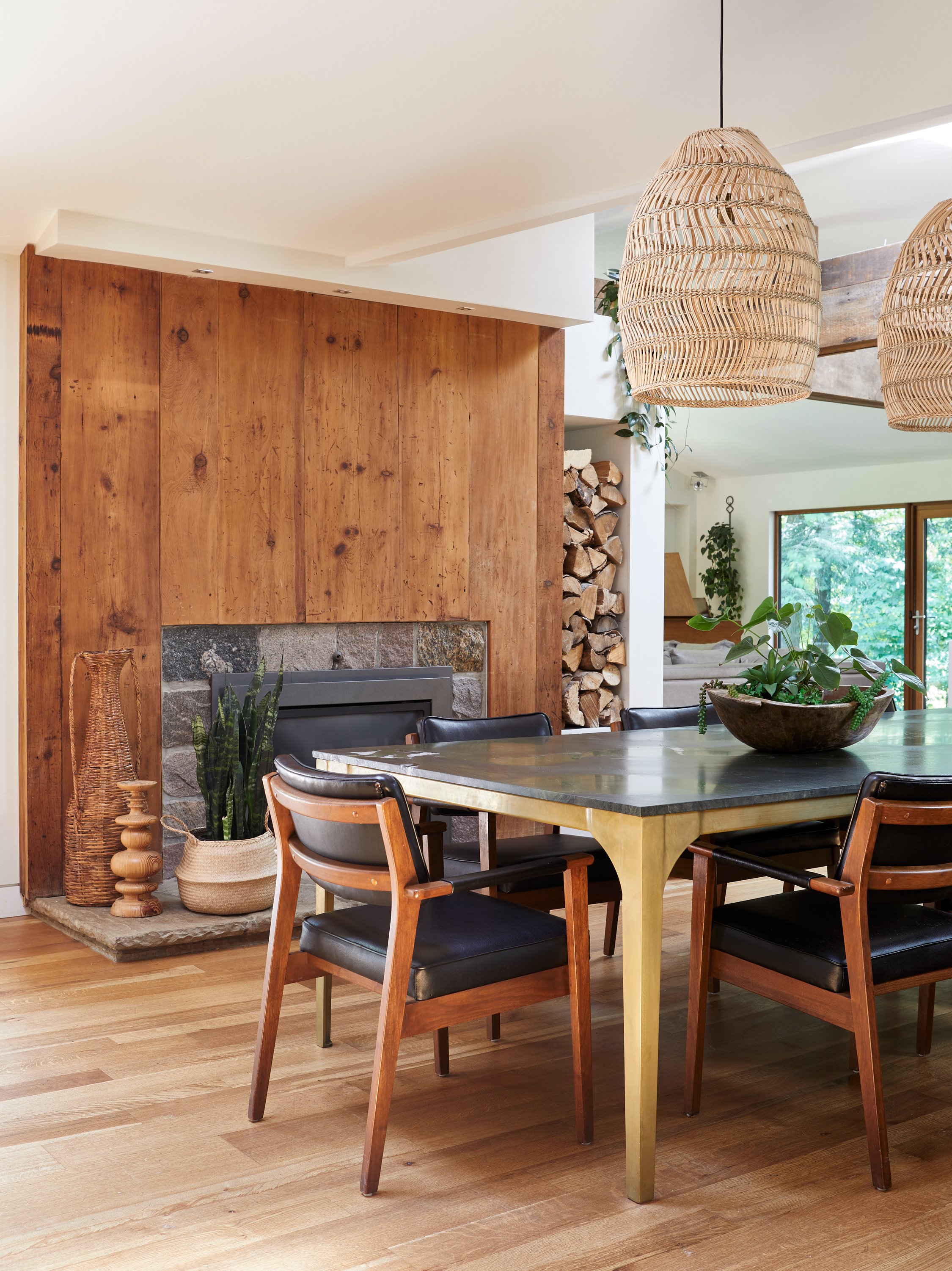 Modern dining room with wooden floor and walls, a stone fireplace, and a large wooden dining table surrounded by black and wood chairs; woven pendant lights hang from the ceiling, and a large bowl with greenery is on the table.