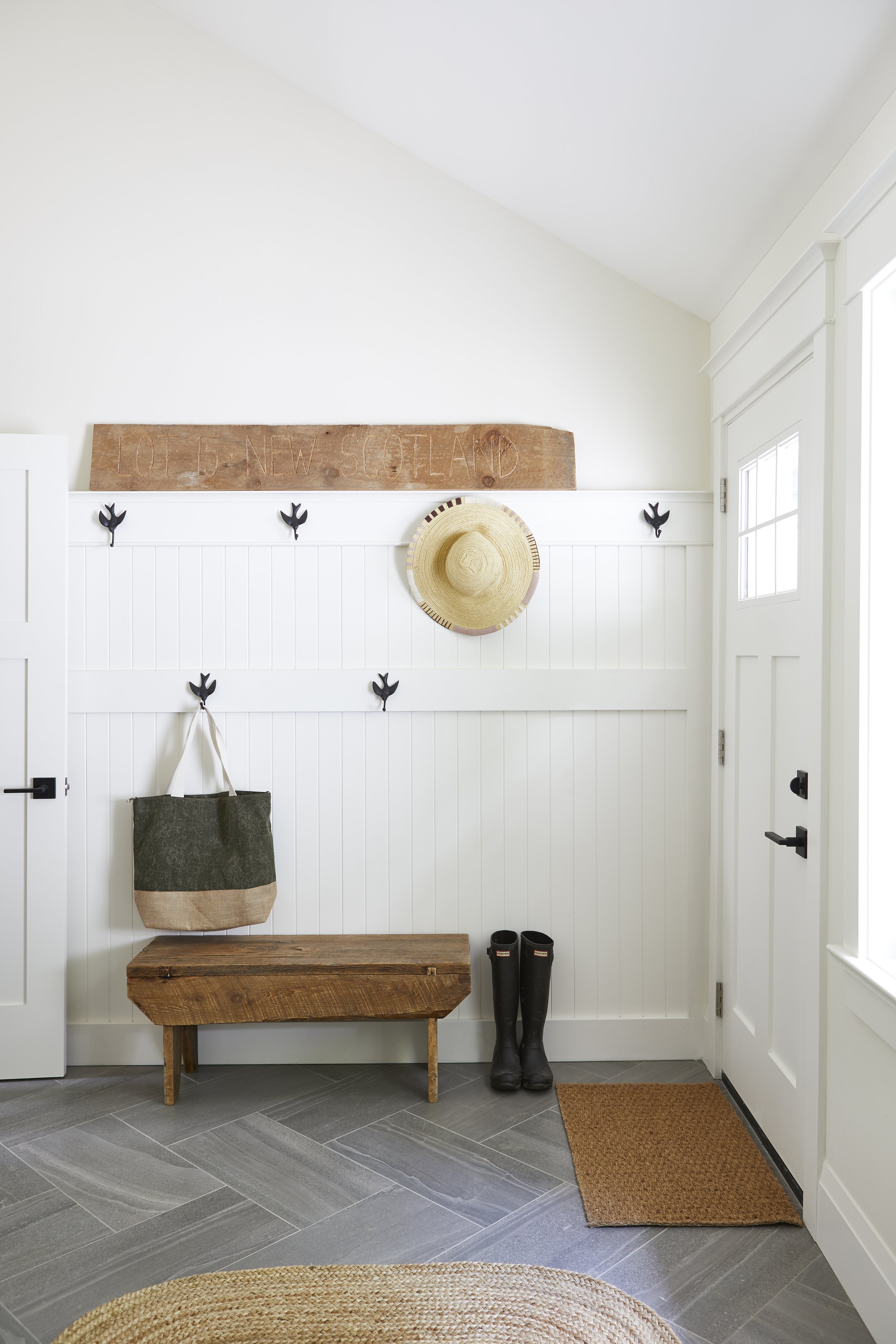 Entryway with white paneled walls, a brown wooden bench, tall black rubber boots, a beige doormat, a beige and black tote bag, a round straw sunhat hanging on a hook, and a wooden plank with 'LOT 45 NEW SCOTLAND' engraved, held on the wall.