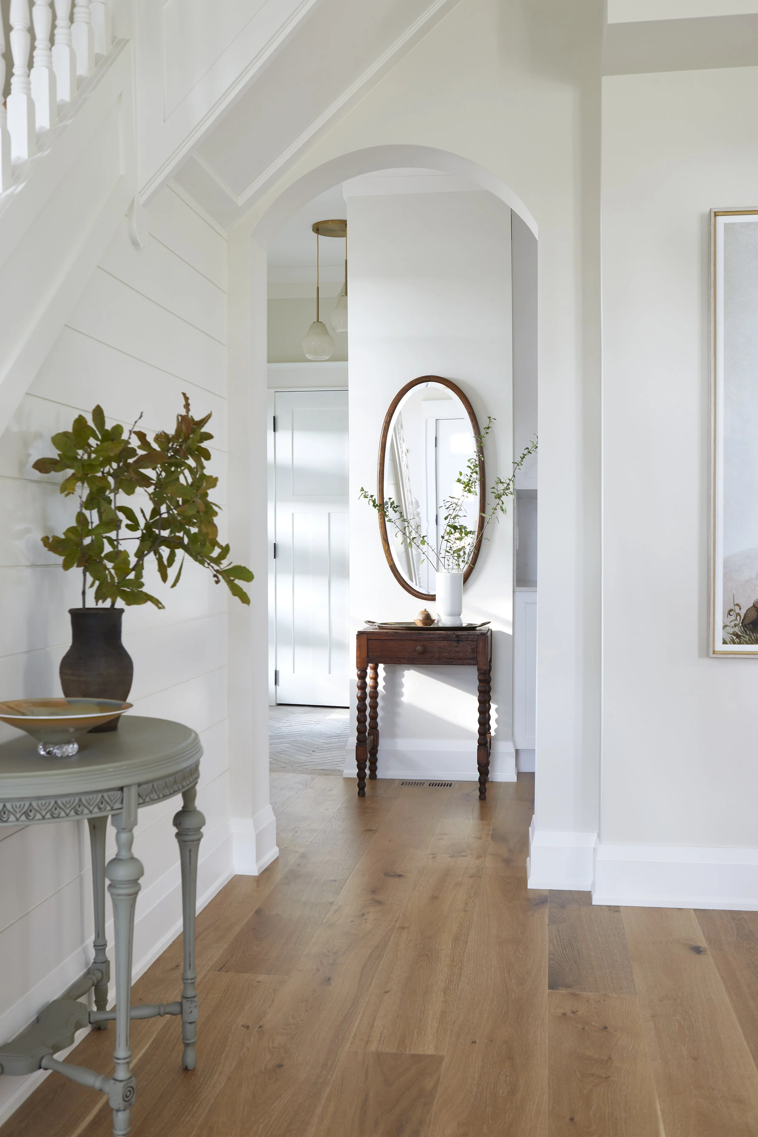 Interior view of a bright, modern home with a wooden floor, white walls, and simple decor including a round side table with a large potted plant, a small wooden table with an oval mirror above it, and a white door in the background.