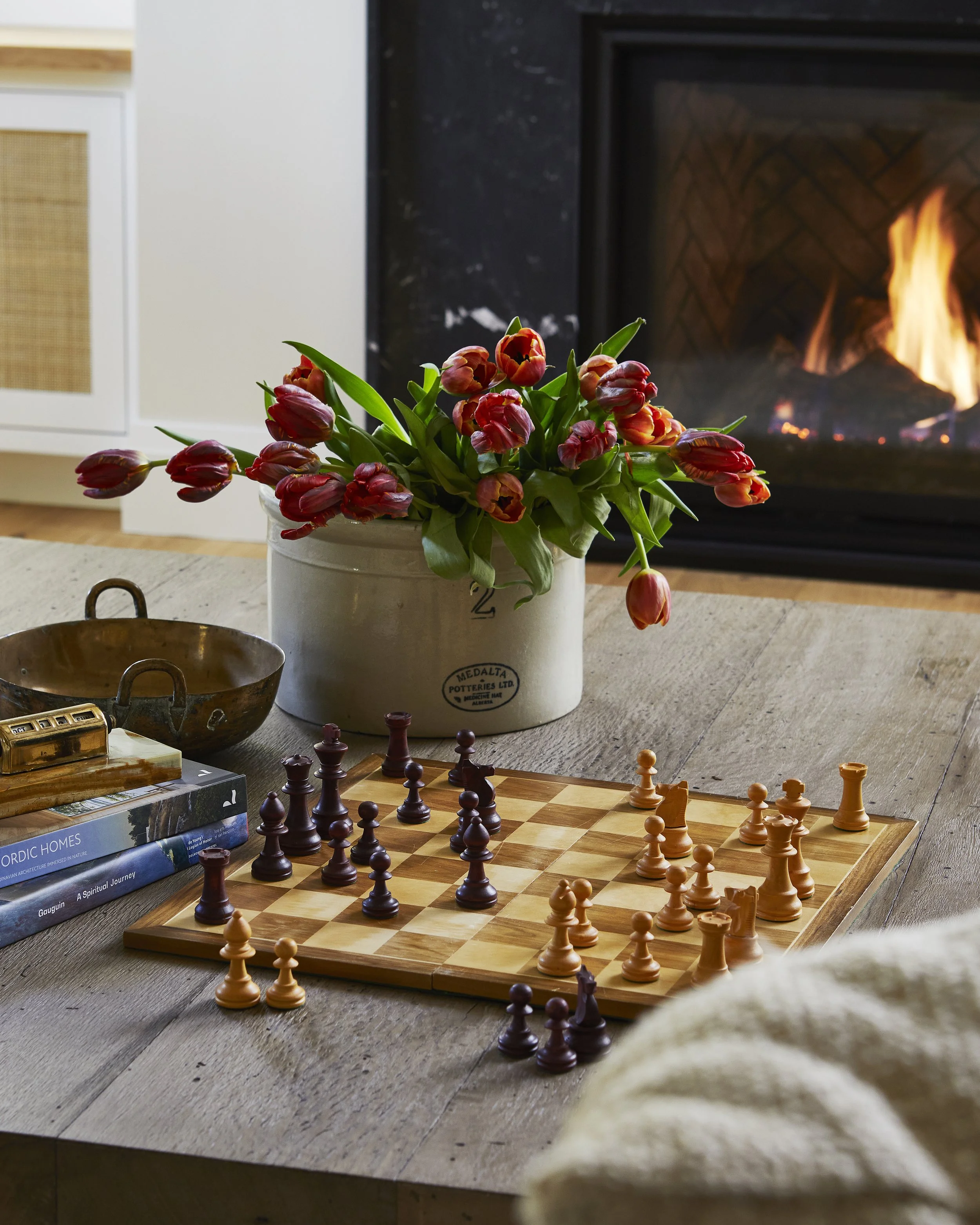A chessboard with pieces arranged for a game, a vase of red tulips, a stone bowl, and books on a wooden table in front of a fireplace with a fire burning.