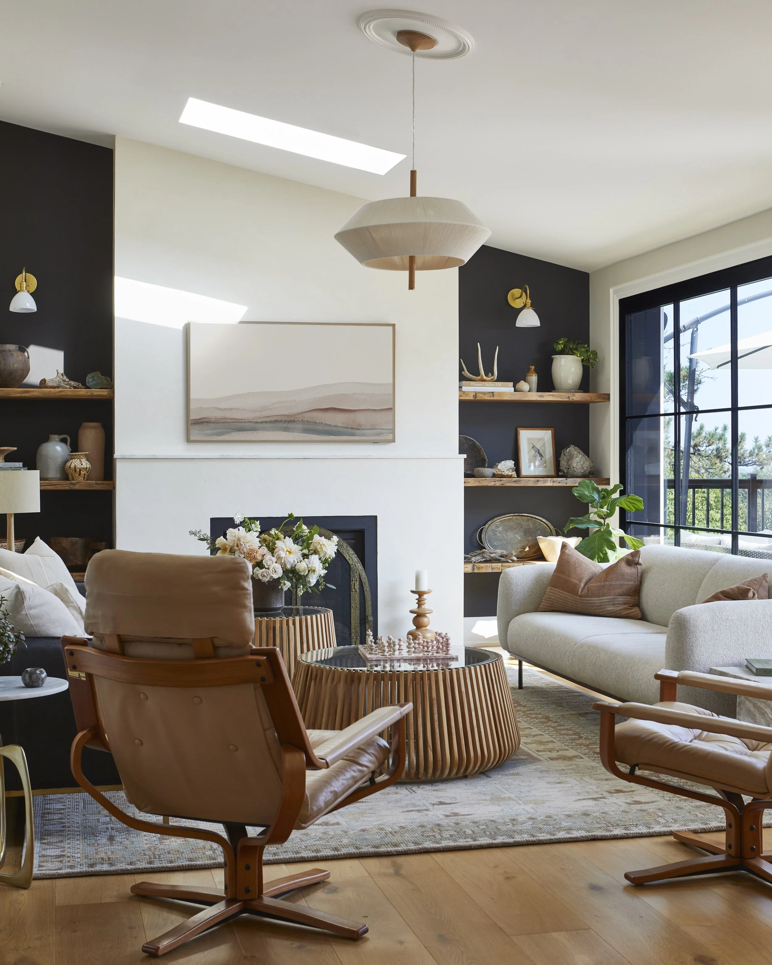 Living room with white sofa, wooden chairs, and a round coffee table, decorated with flowers and candles, with built-in shelves and large sliding glass door leading outside.