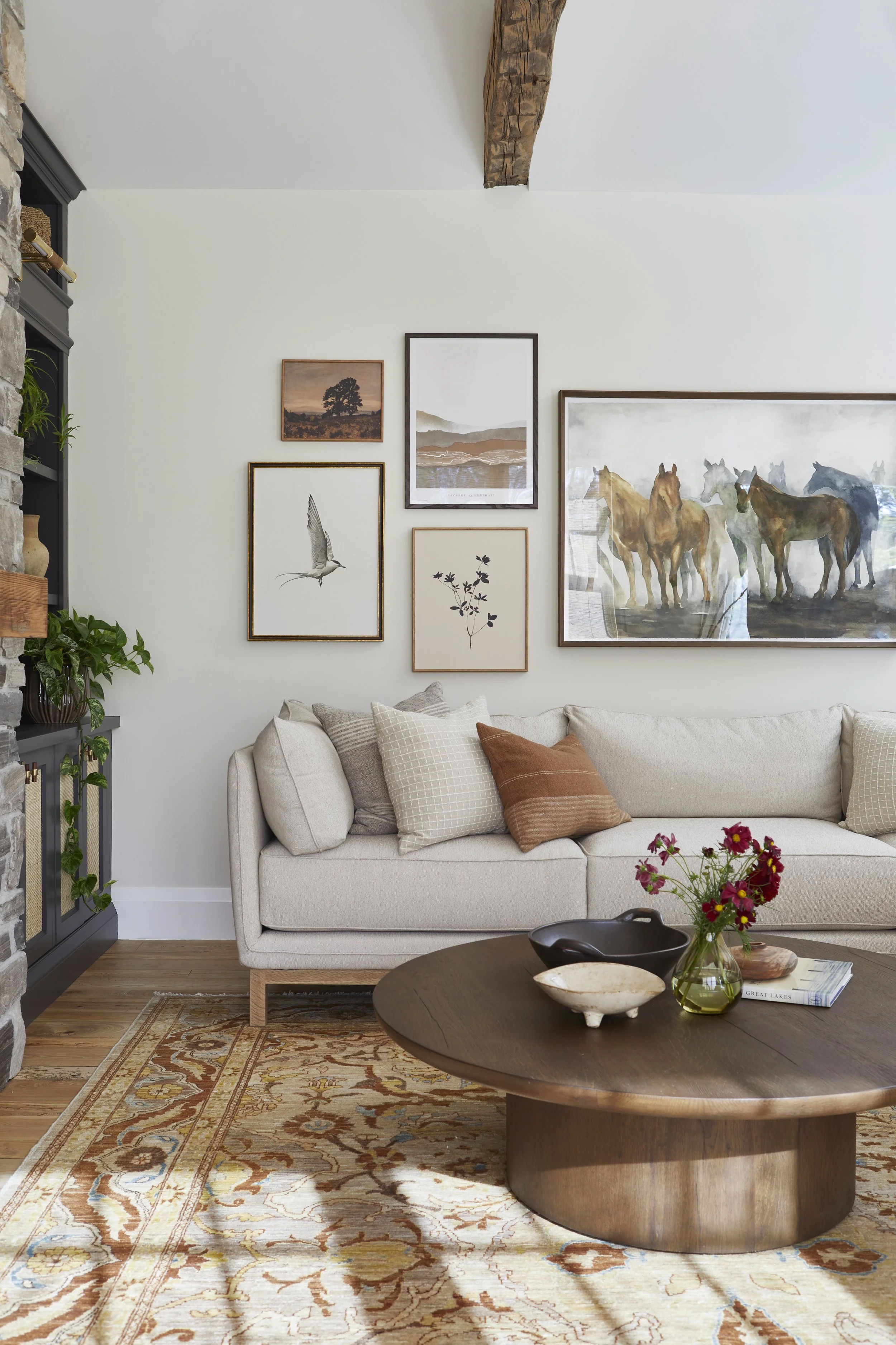 Living room with a beige sofa, framed artwork including horse paintings on the wall, a round wooden coffee table with decorative bowls and flowers, a patterned rug, and a stone fireplace with plants.