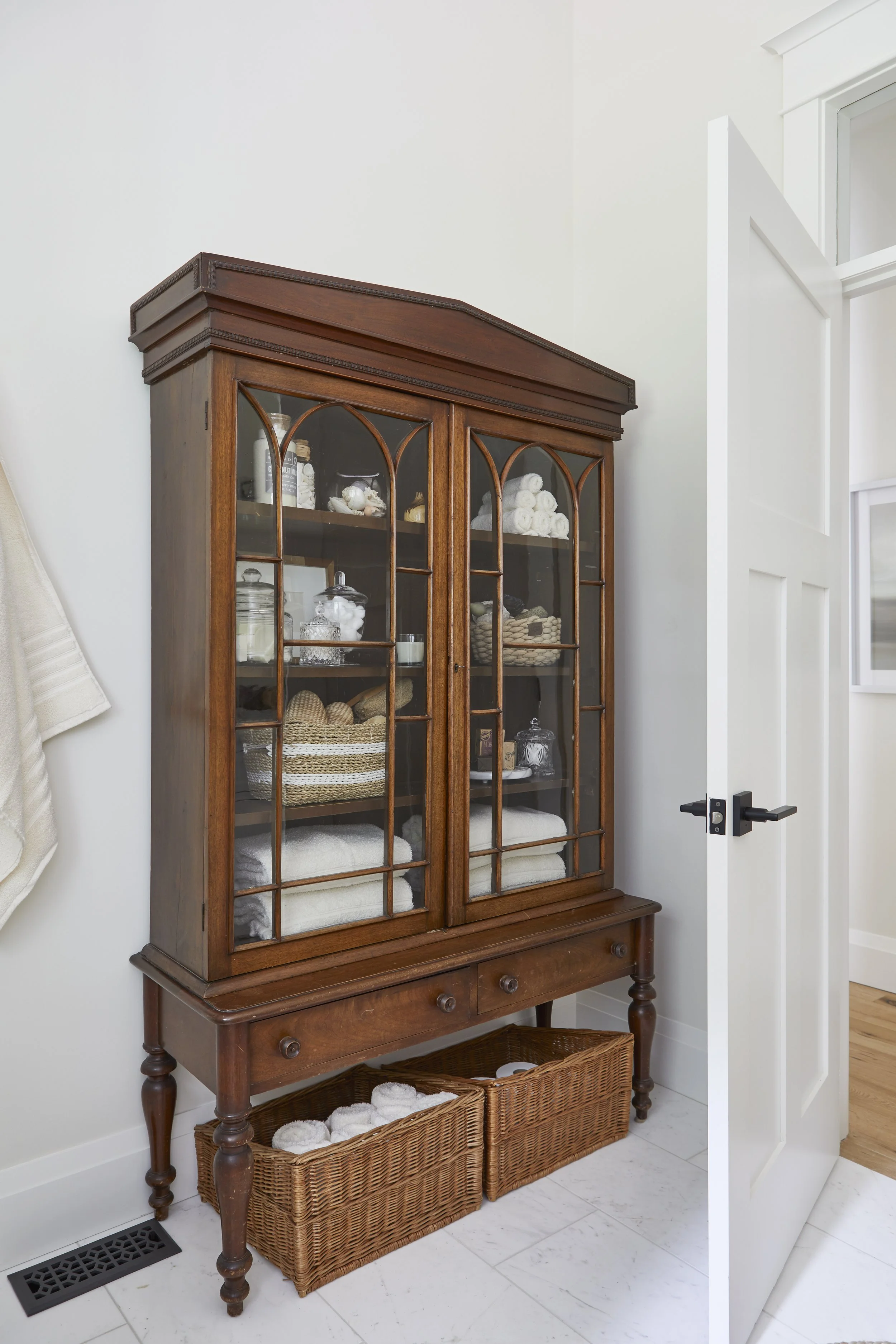 A wooden glass-front cabinet filled with folded towels, toiletries, and decorative items. Two wicker baskets are placed underneath on a lower shelf. The cabinet is next to a white door and on a white wall floor near a floor vent.