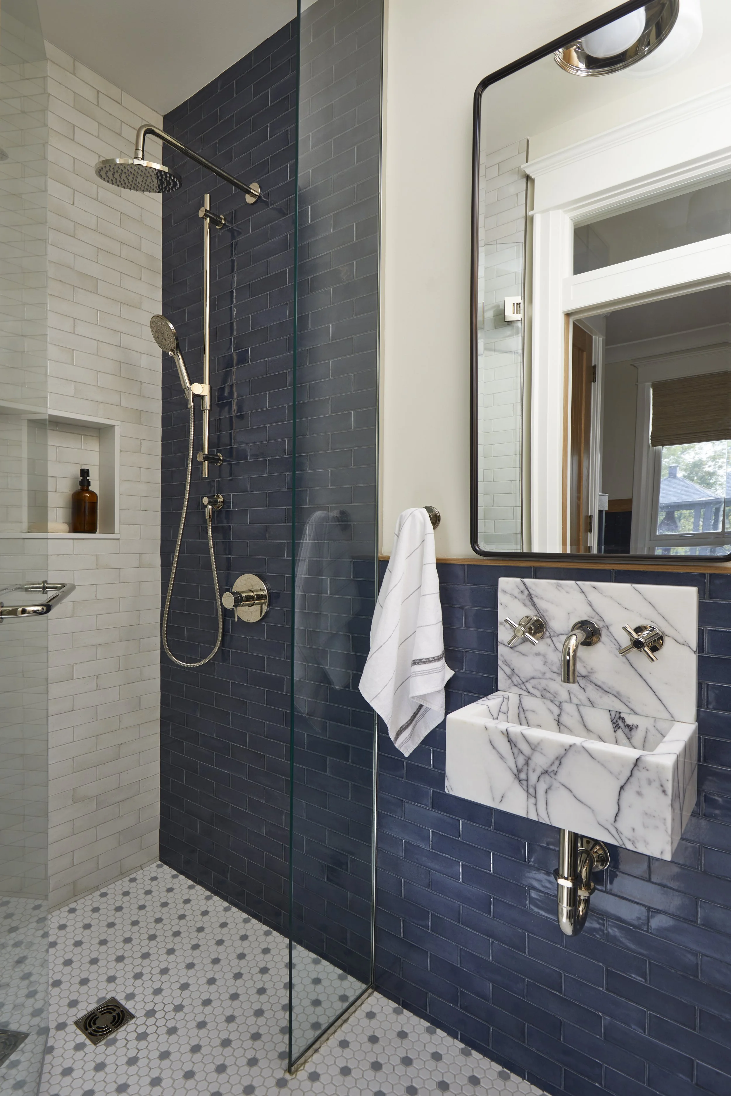 Modern bathroom with a walk-in shower featuring a dark blue tiled wall, a glass partition, and a white tiled wall inside the shower. There is a marble sink with vintage-style fixtures, a large mirror above, and a window with a brown blind in the back