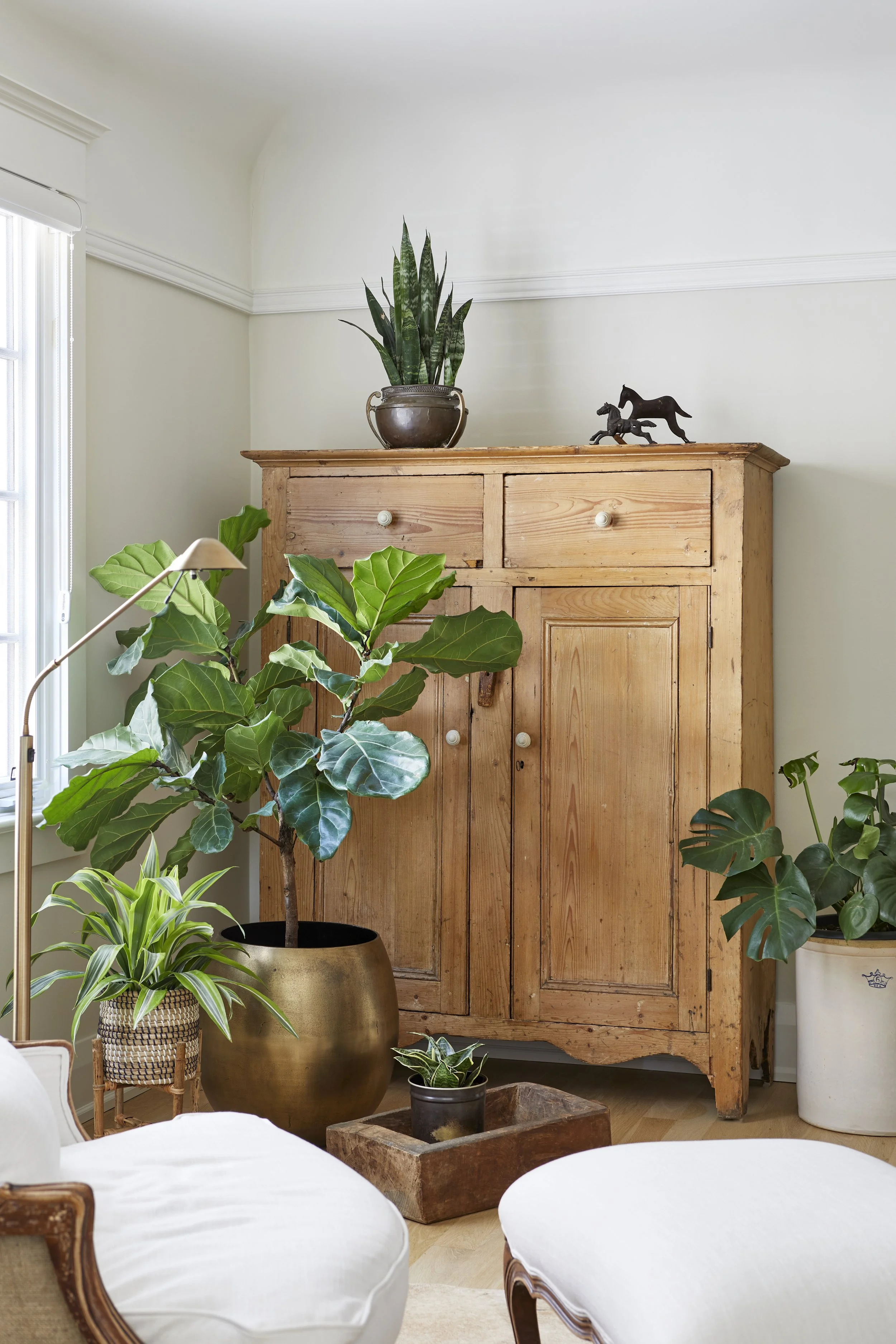 Living room corner with wooden cabinet, potted green plants, a table lamp, and decorative horse sculptures.