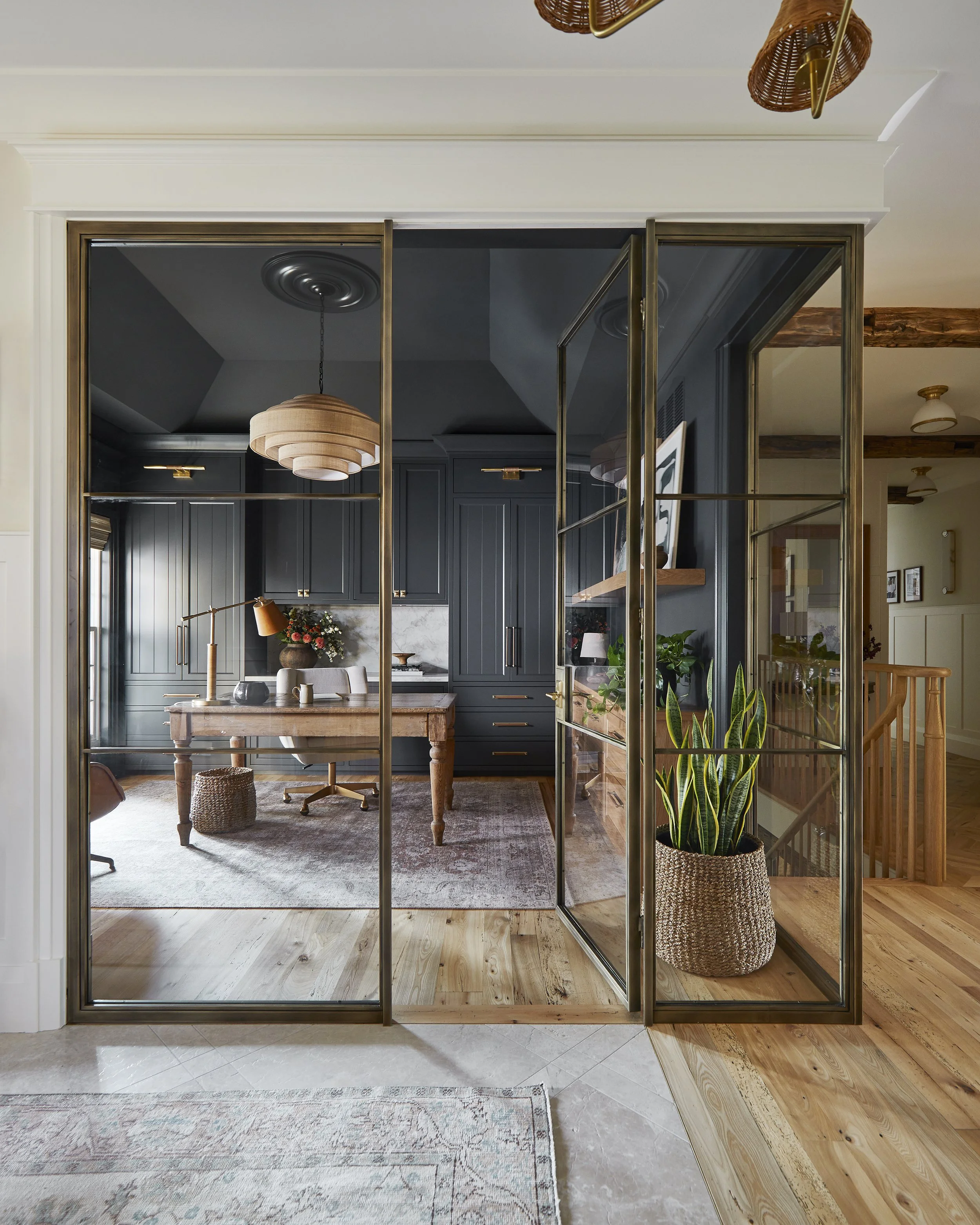 Modern home office with dark blue cabinetry, wooden desk, potted plant, and gold-framed glass doors.