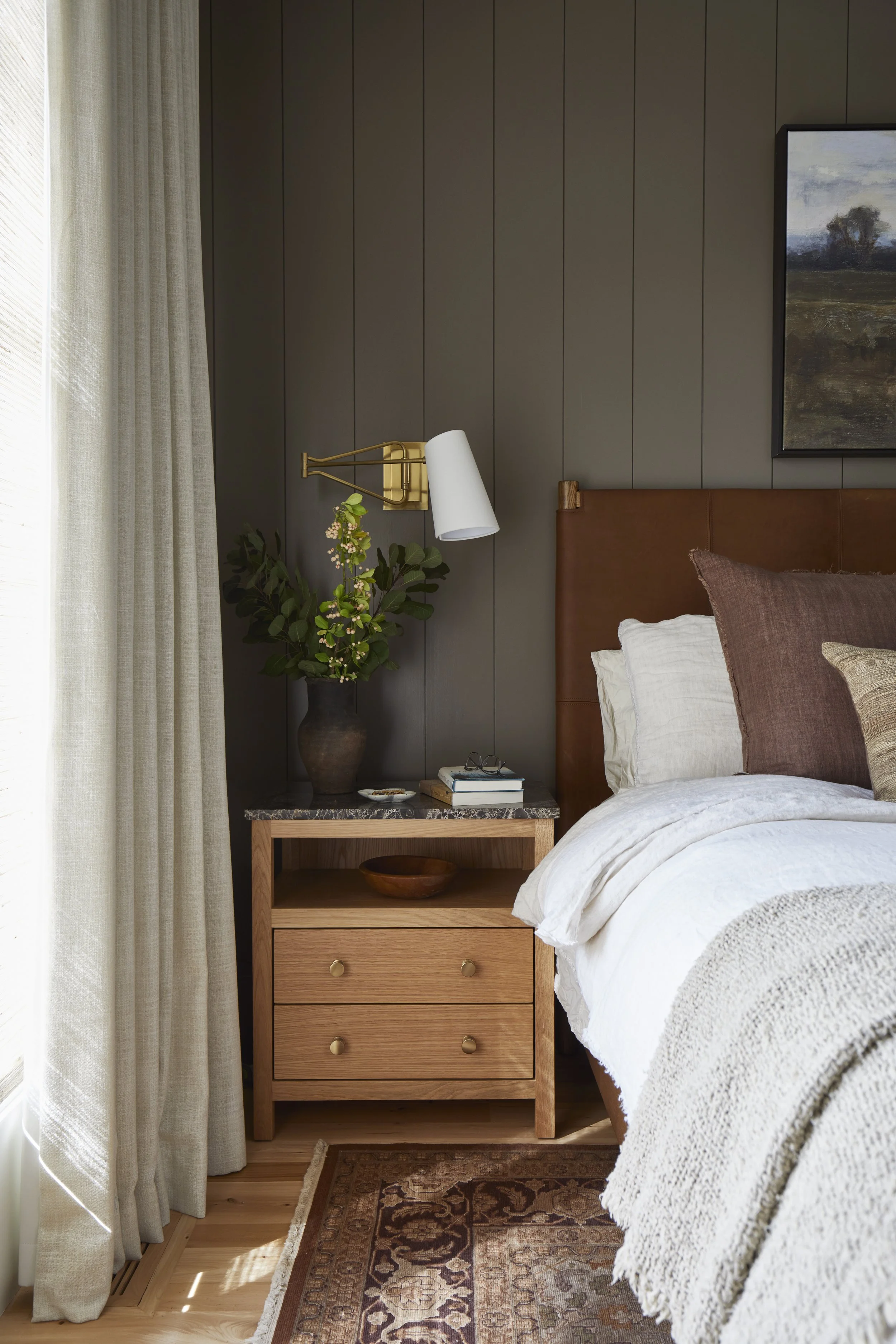 A cozy bedroom with a beige nightstand on the left side of the bed, holding a dark vase with green foliage, a small dish, and a couple of books. A wall-mounted brass and white adjustable reading lamp is above the nightstand. The bed has a brown leath