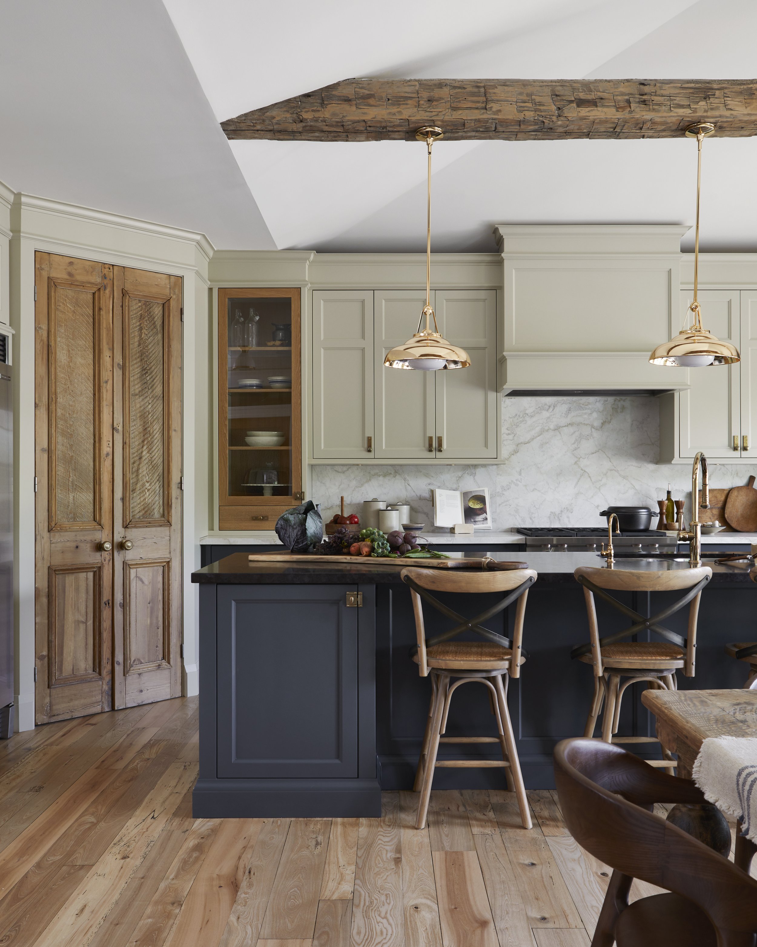 Kitchen with light gray cabinets, a dark gray island, wooden chairs, and wooden floors. There are two gold pendant lights and a rustic wooden beam on the ceiling.