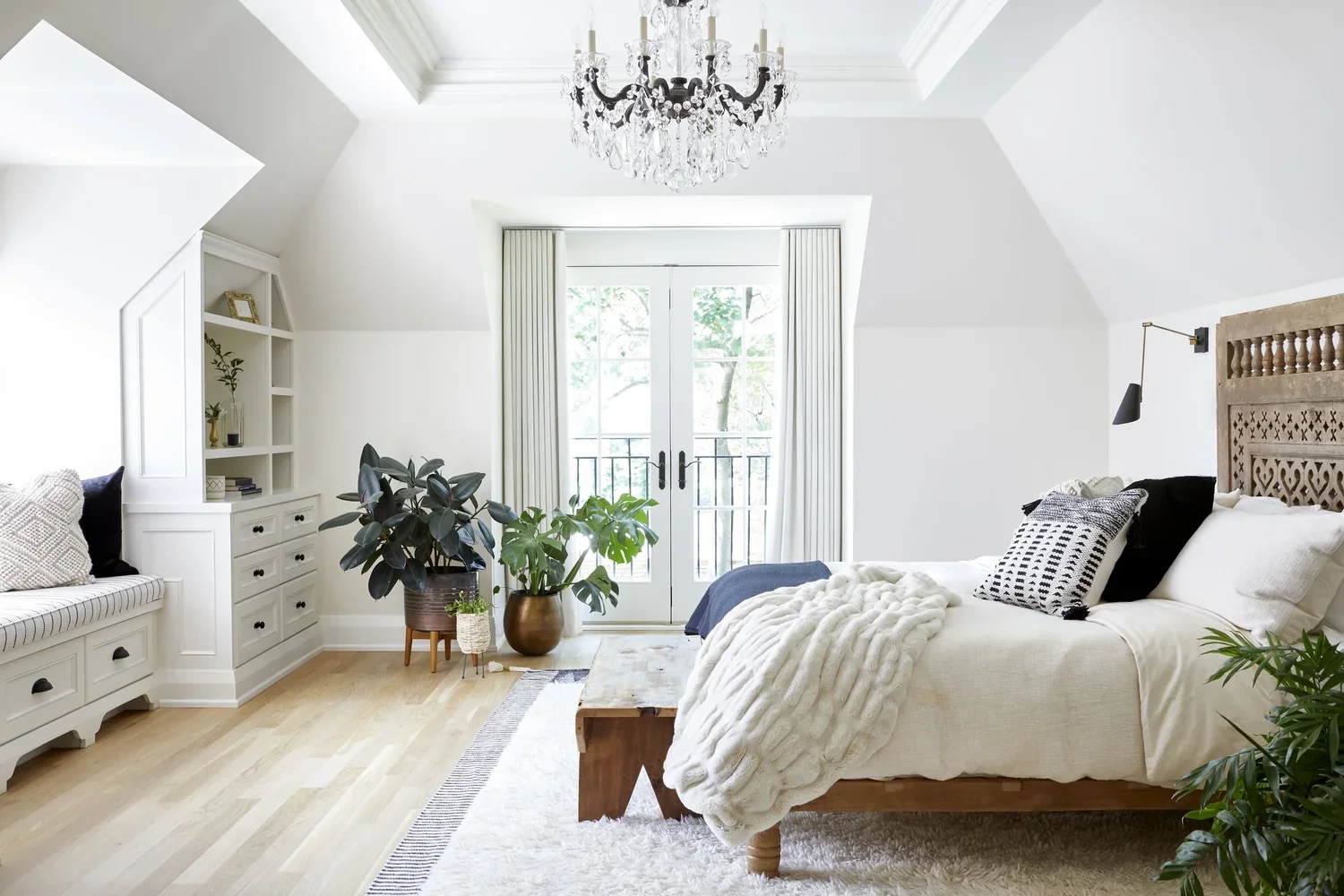 Bright bedroom with white walls, large window with balcony door, wood floor, white bed with black and white pillows, cozy blanket, bedside wall lamp, large potted plants, built-in white cabinet with open shelves, chandelier, and a wooden bench at the