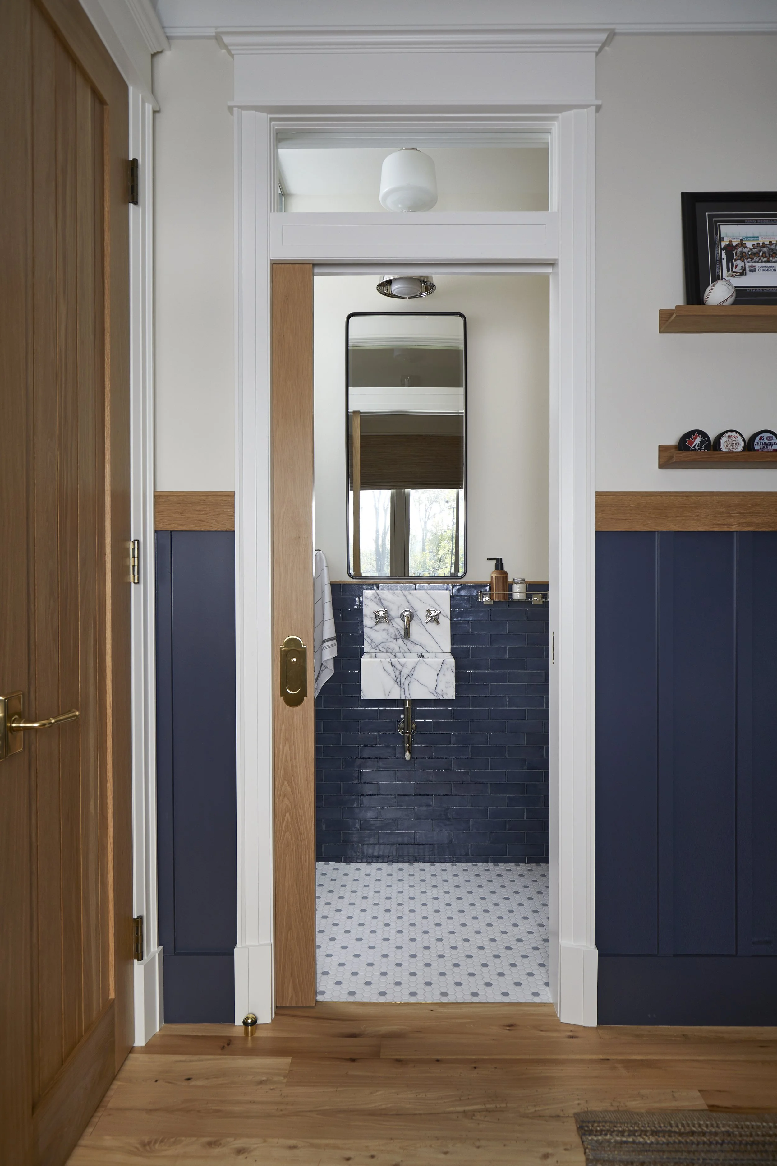 View of a small bathroom with a marble sink, dark navy blue tiled wall, and a mirror, seen through a doorway with white trim. The bathroom has wood accents and a small window reflecting greenery outside.