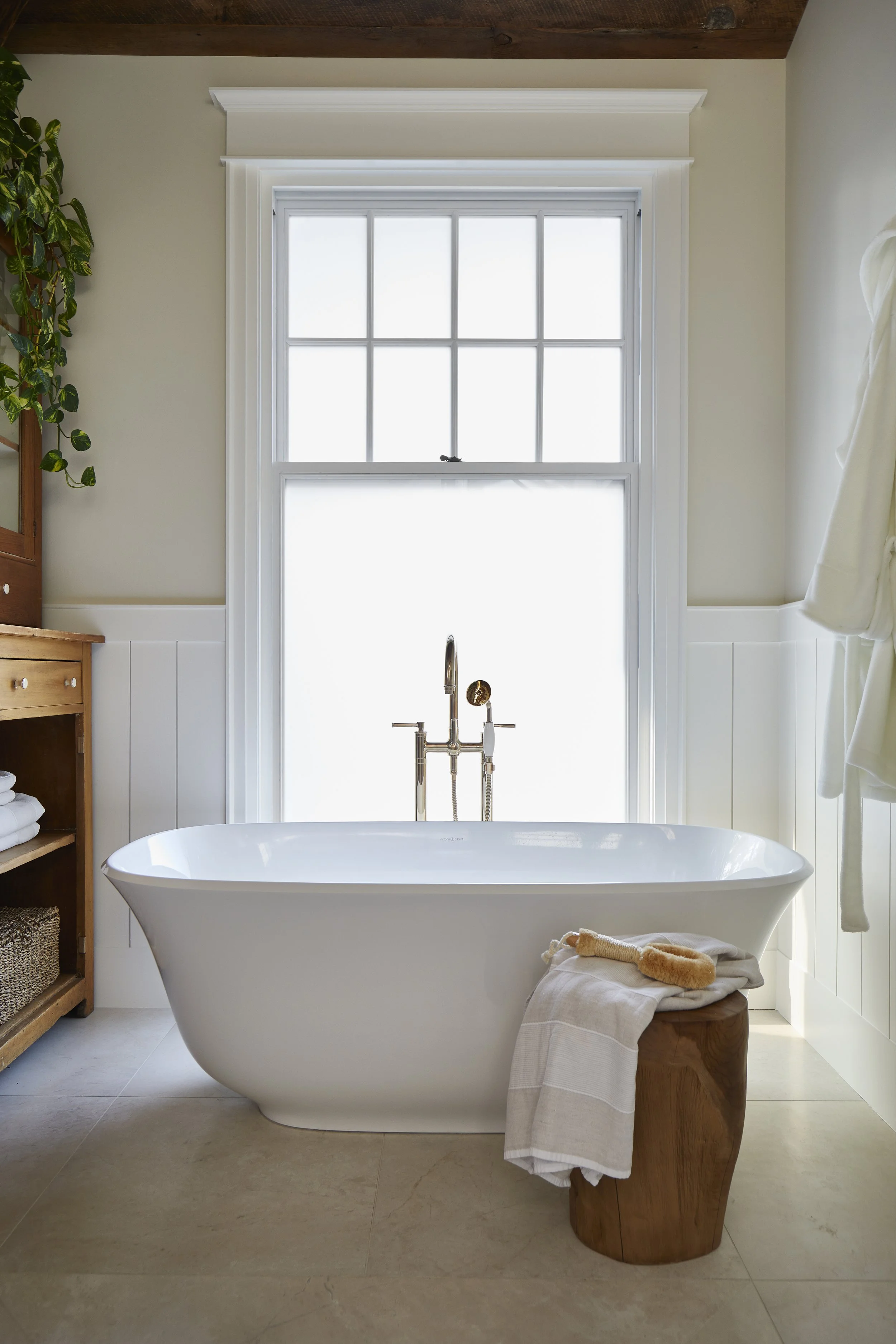 A modern bathroom featuring a standalone bathtub in front of a large window with frosted glass, with towels and a bath brush draped over a small wooden stool.
