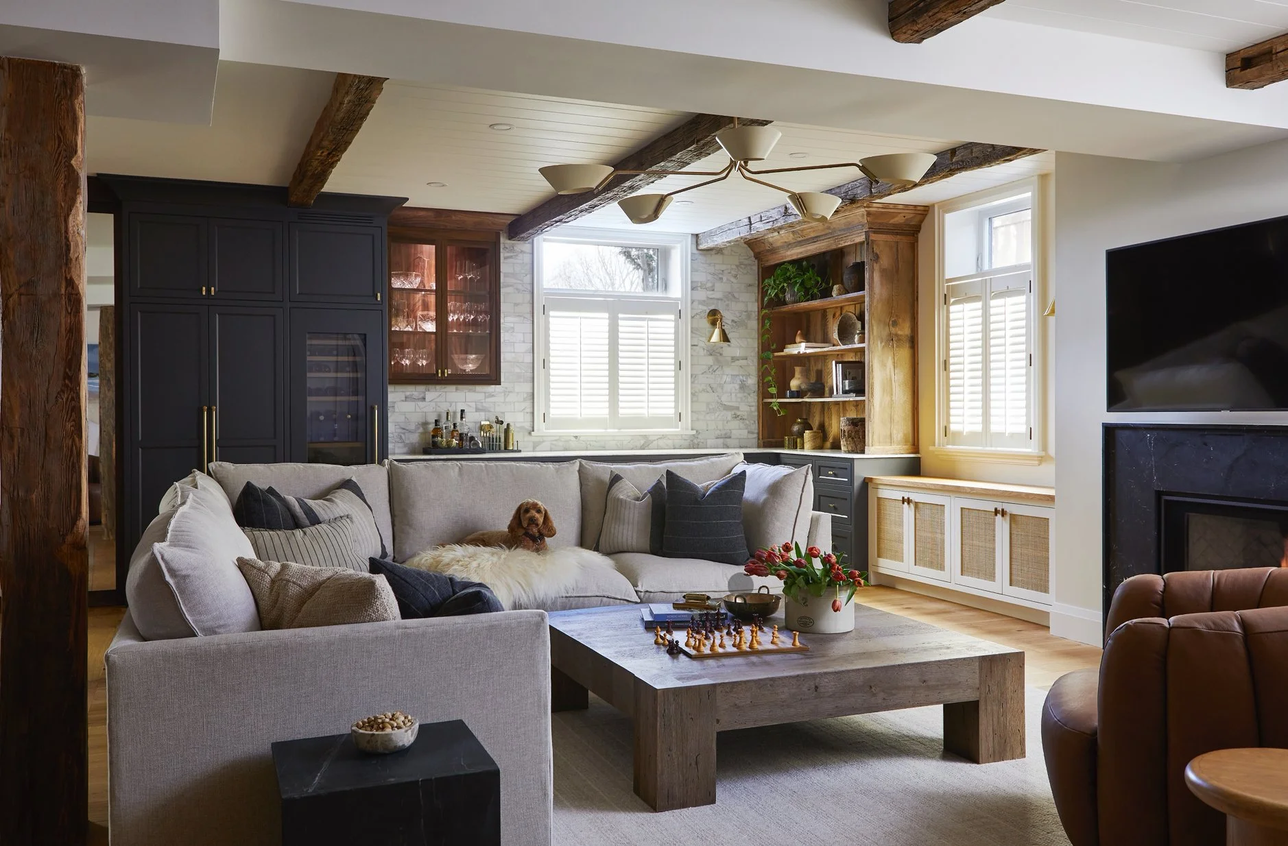 Living room with a beige sectional sofa, wooden coffee table with a chessboard, and a small dog lying on the sofa. The room has a mix of natural and rustic decor, with dark and light cabinetry, open shelves, and a fireplace with a television above. W