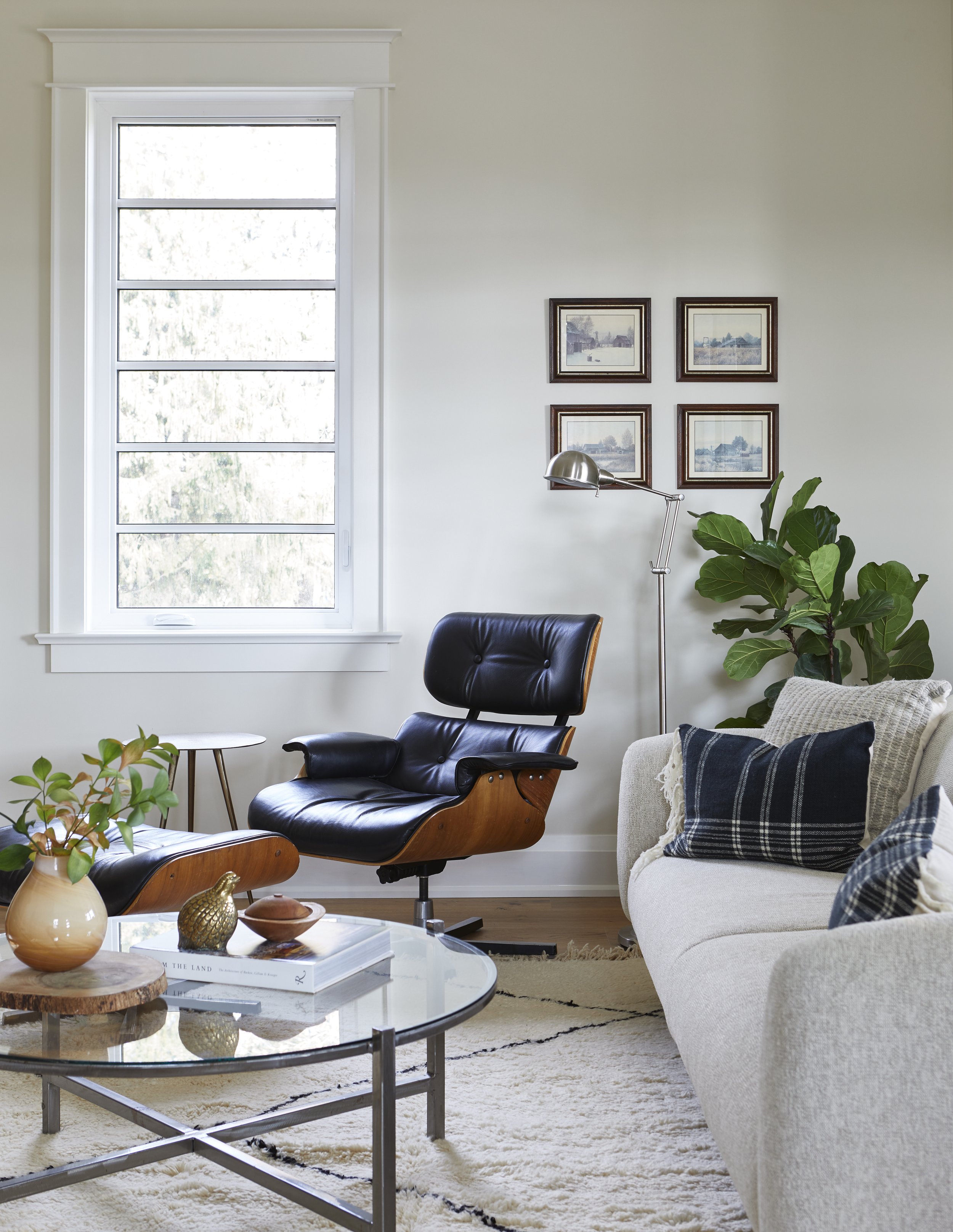 Living room with a black leather and wood lounge chair, a beige sofa with plaid pillows, a round glass coffee table with decorative items, a window with white trim, framed artwork on the wall, a green plant, and a floor lamp.