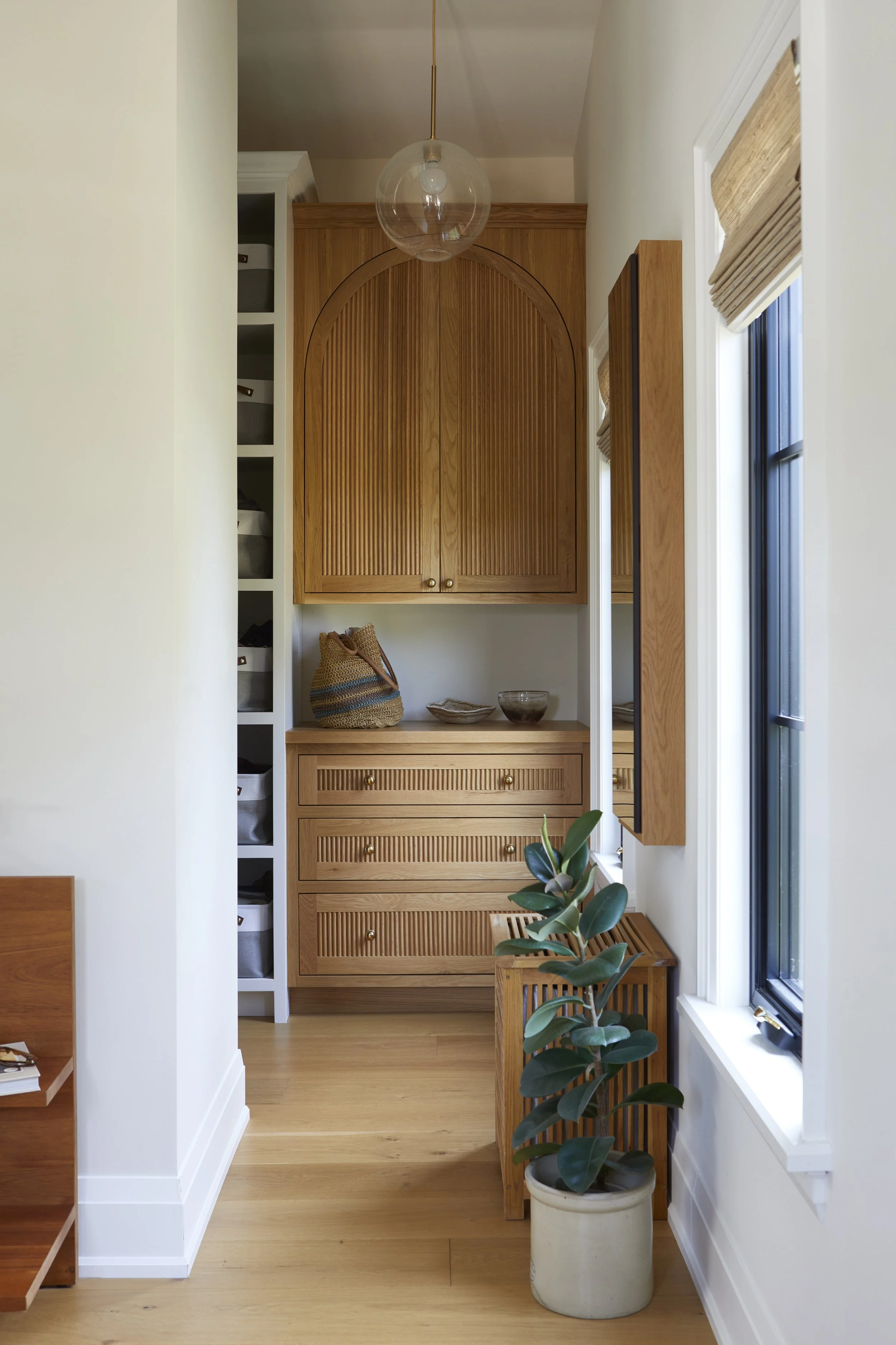 Interior of a room with wooden furniture, a potted plant, a window with a beige Roman shade, and a hanging glass light fixture.