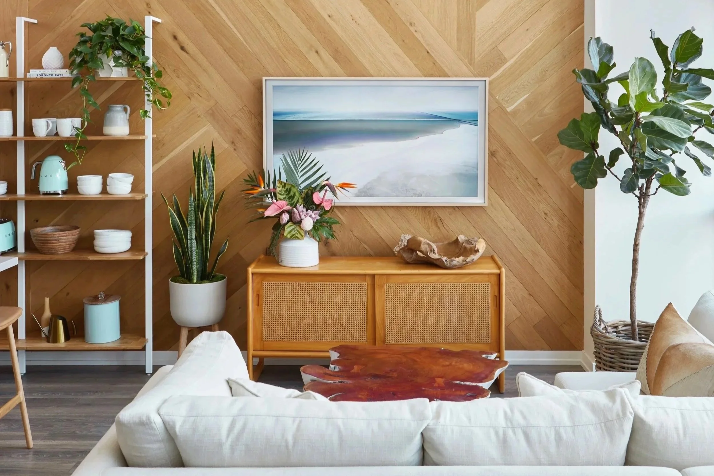Living room with a beige sofa, a wooden coffee table, a light wood sideboard, a framed ocean beach photograph, potted plants including a snake plant and a fiddle leaf fig, an open wood and white shelf with ceramics and a plant, and a wood-paneled accent wall.