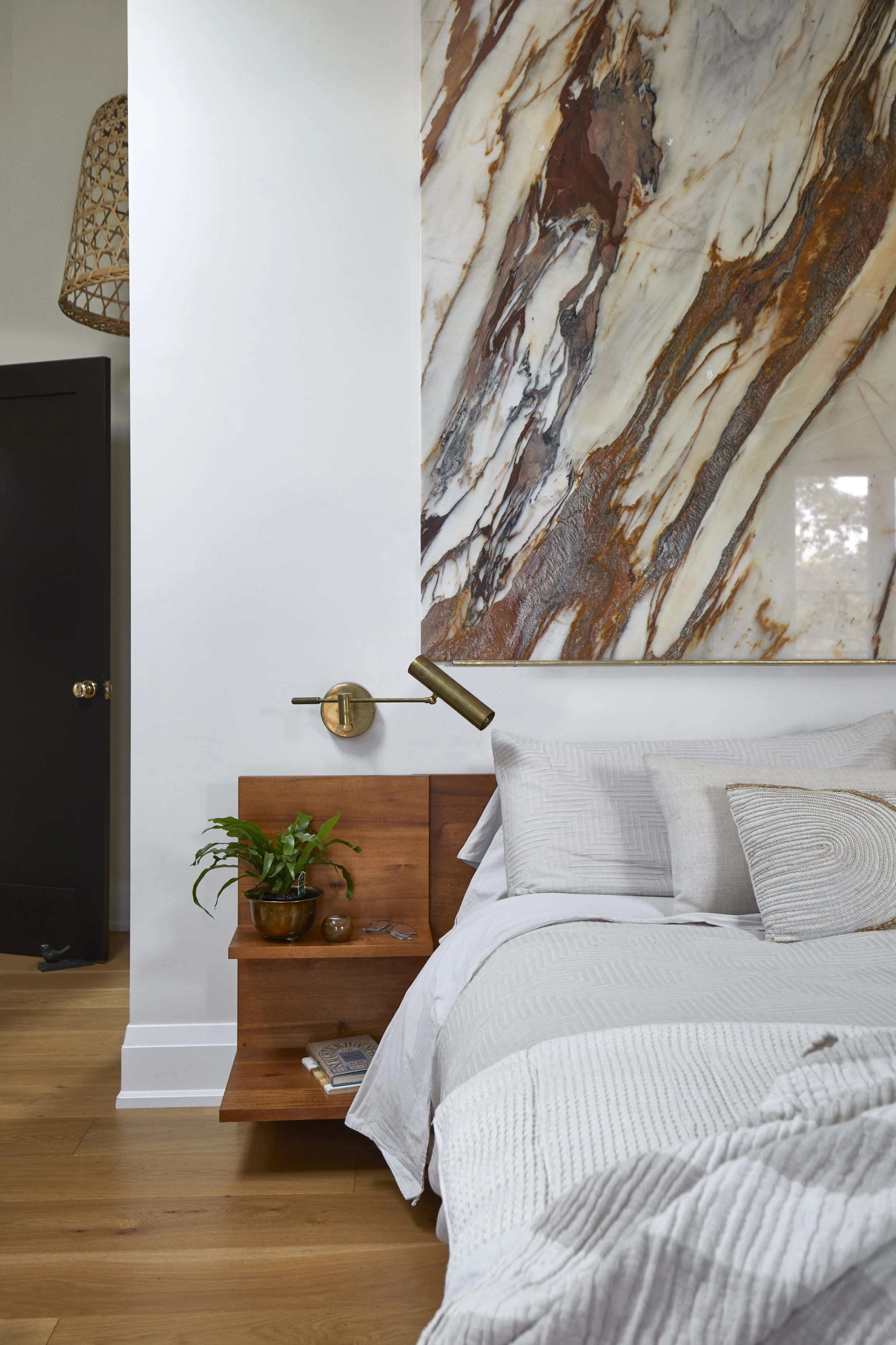 A bedroom corner with a bed, wooden headboard, and a built-in nightstand holding a small potted plant, a book, and a pair of glasses. A brass wall lamp extends over the bed, and a large abstract marble artwork hangs above the bed. Part of a black war