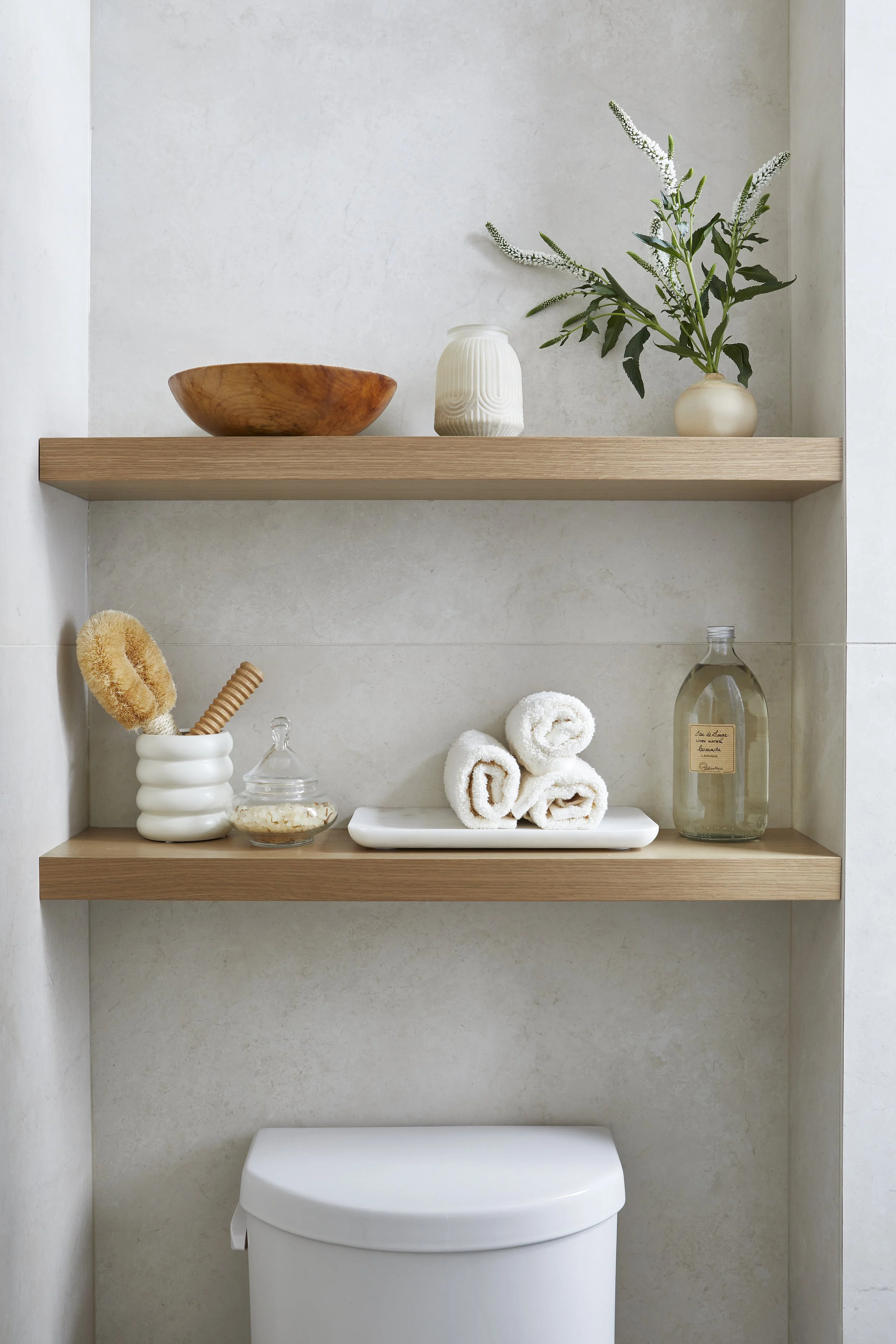 Decorative bathroom shelves with a wooden bowl, ceramic vase with green leafy branches, rolled white towels, a glass jar, a bottle of liquid soap, and spa accessories.