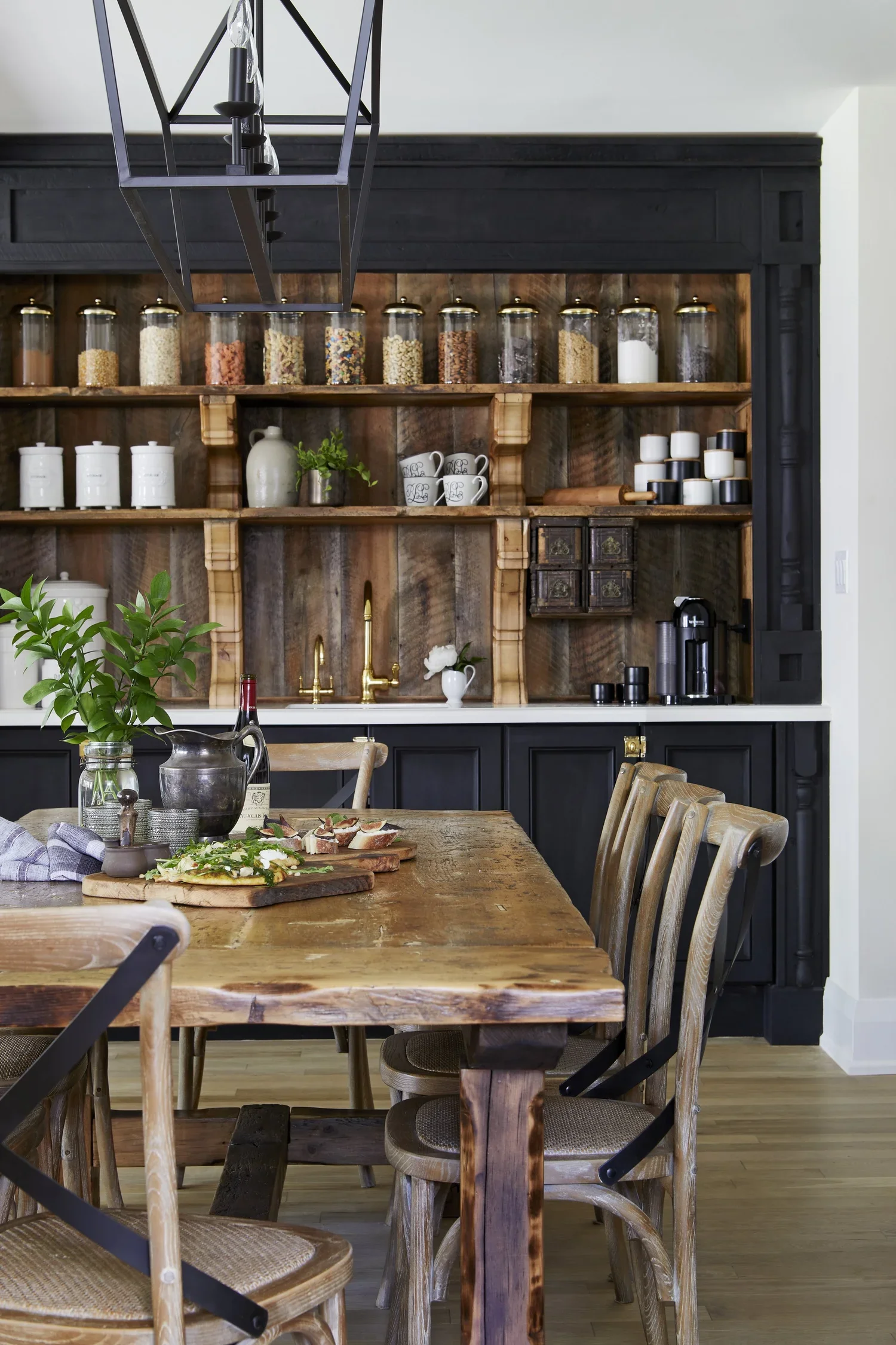 A rustic kitchen and dining area featuring a wooden table with chairs, greenery, and a wall-mounted open shelving unit with jars, cups, and decorative items. A black chandelier hangs above the table.