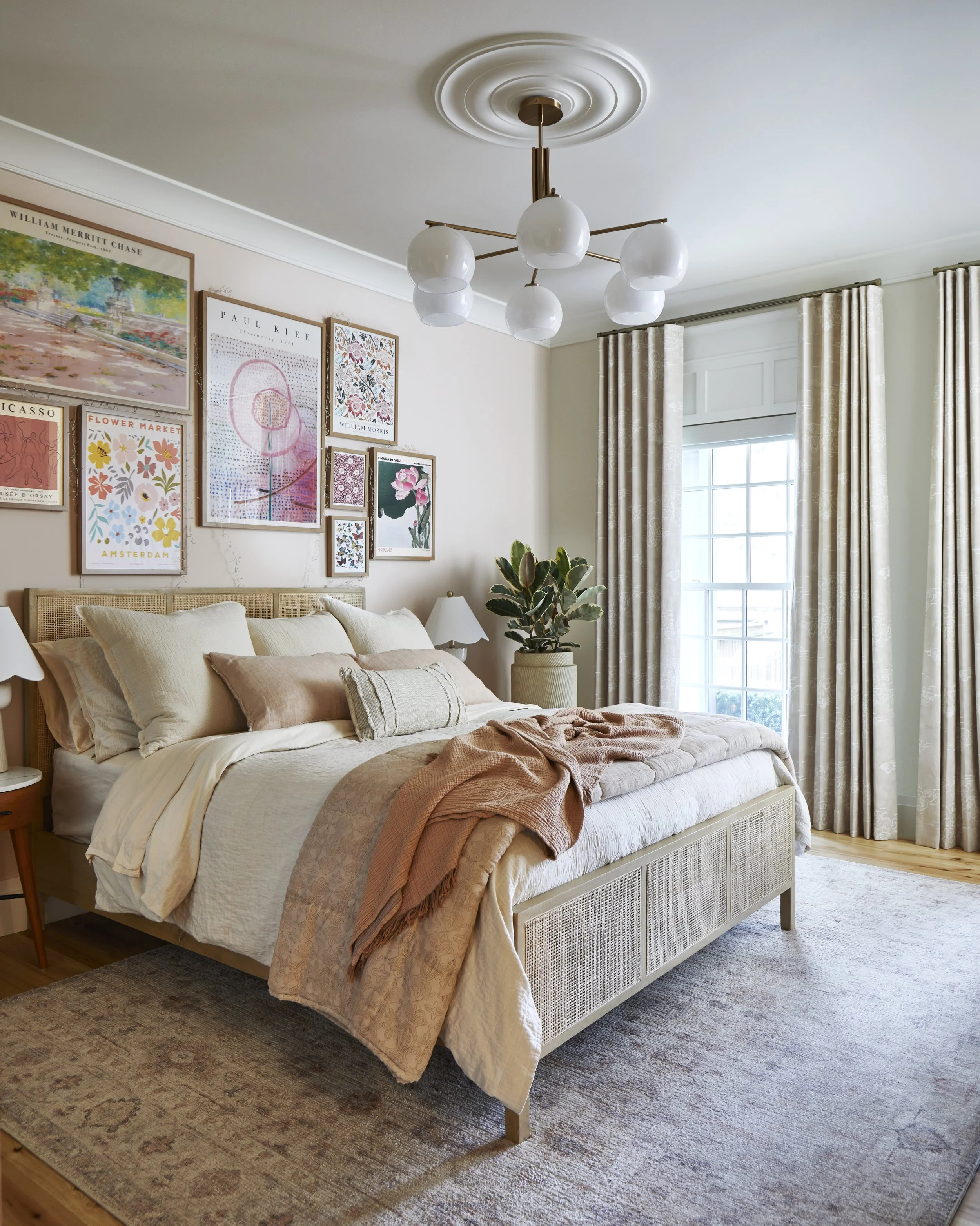 A cozy bedroom with a beige bed adorned with multiple pillows and peach-colored throws, a side table with a lamp, large window with patterned curtains, a potted plant, and a gallery wall of colorful art prints above the bed, illuminated by a modern c