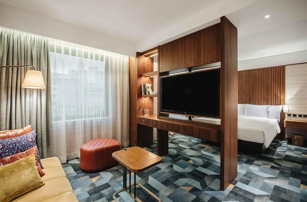 Chambre d'hôtel moderne avec un espace salon séparé par une cloison en bois, grande fenêtre avec rideaux, lit king-size dans la chambre principale, canapé avec coussins, bureau en bois, et une télévision à écran plat.