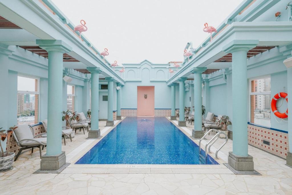 Piscine intérieure avec des colonnes bleues, chaises longues avec des coussins, fenêtres offrant une vue urbaine, un dispositif de sauvetage en anneau à la paroi, décor de flamants en plastique sur la rambarde.