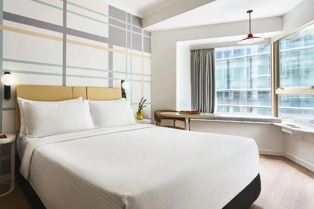 Chambre d'hôtel moderne avec lit double, grand fenêtre avec vue sur des immeubles, mur mural graphique, table de travail, lampes de lecture, et décoration minimaliste.