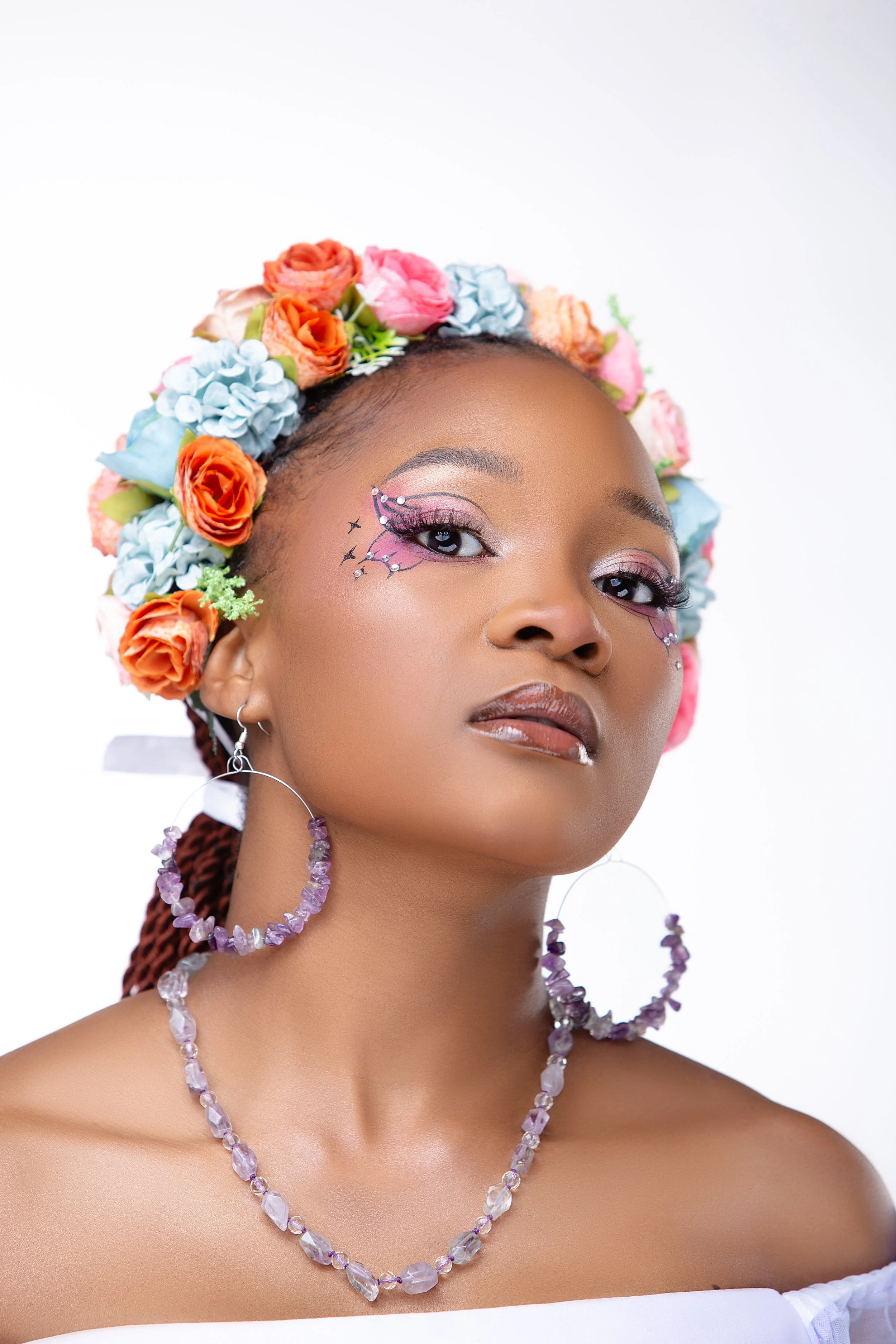 A woman wearing a floral crown with pink, orange, and blue flowers, makeup with pink and purple eyeshadow, and jewelry including a purple beaded necklace, earrings, and a facial piercing.