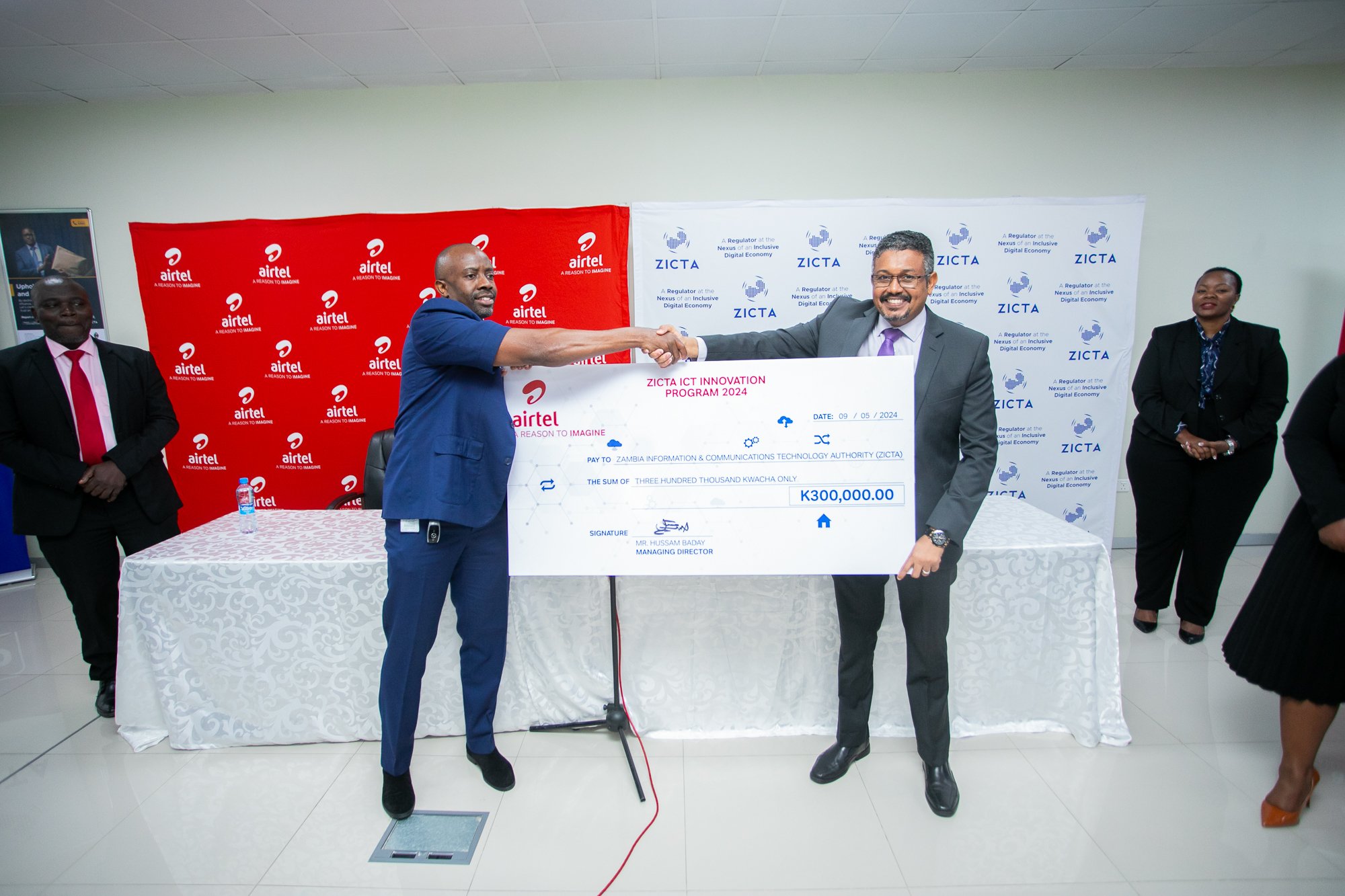 Two men shaking hands at a formal event, holding a large ceremonial check made out to the Zambia Information & Communications Technology Authority for 300,000 kwacha, with three other individuals standing nearby. Background features banners from Airt