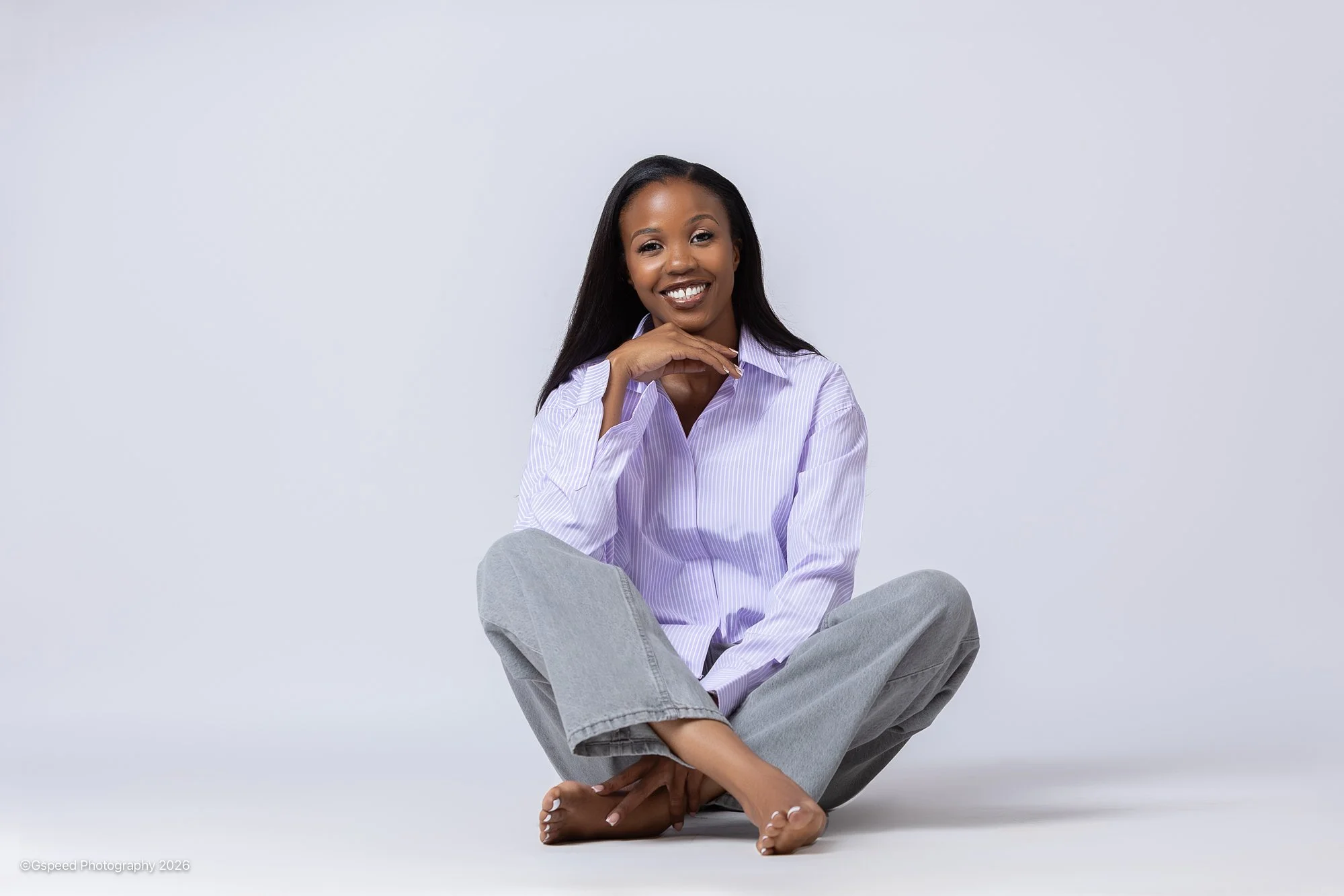 A woman sitting cross-legged on the floor against a plain light gray background, smiling and resting her chin on her hand. She is wearing a light purple striped button-up shirt and gray pants.