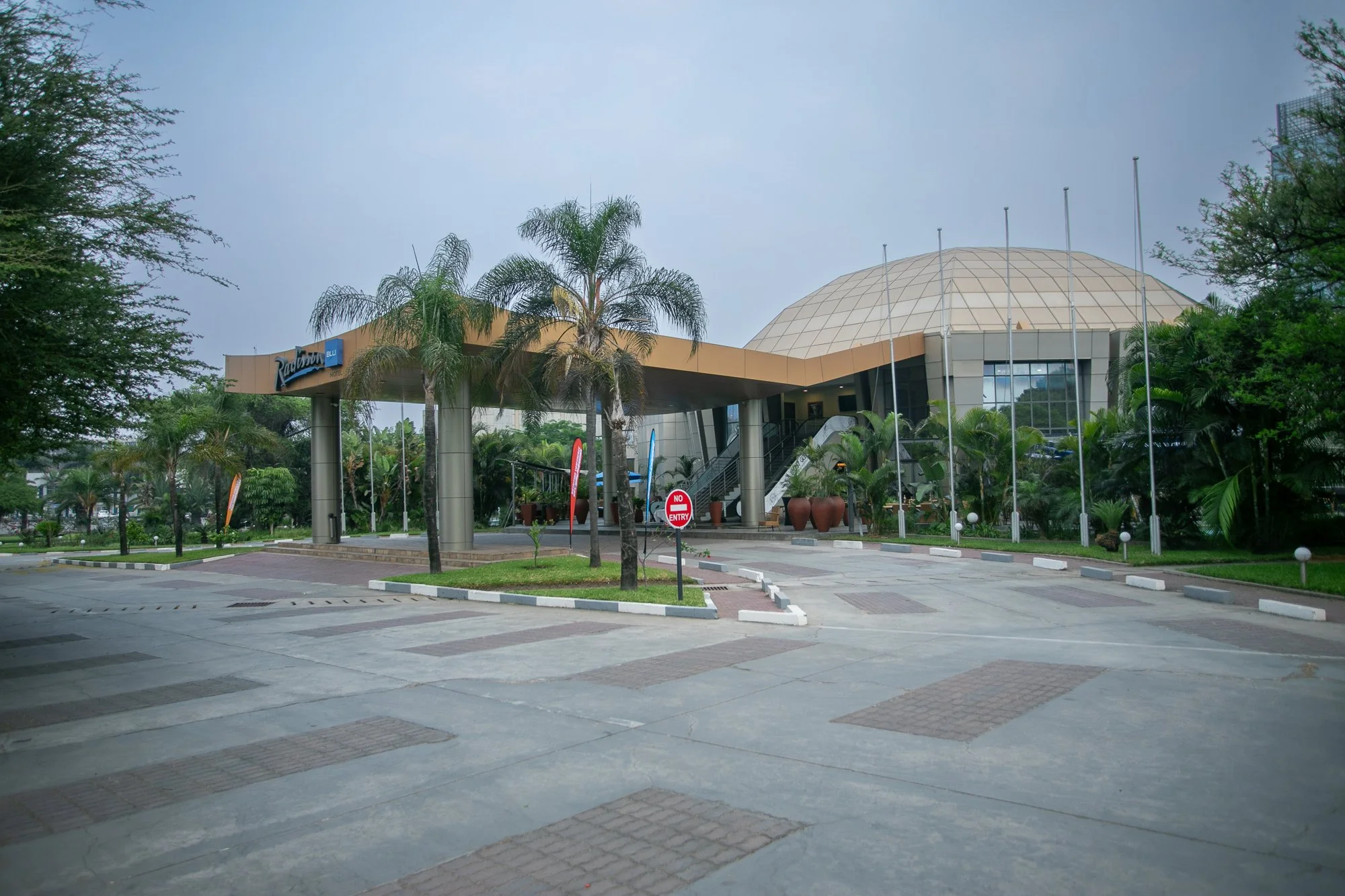 The image depicts a modern building with a large dome structure on the right side and a flat roof on the left side, surrounded by trees and palm trees. There are flagpoles and a parking area in the foreground, along with 