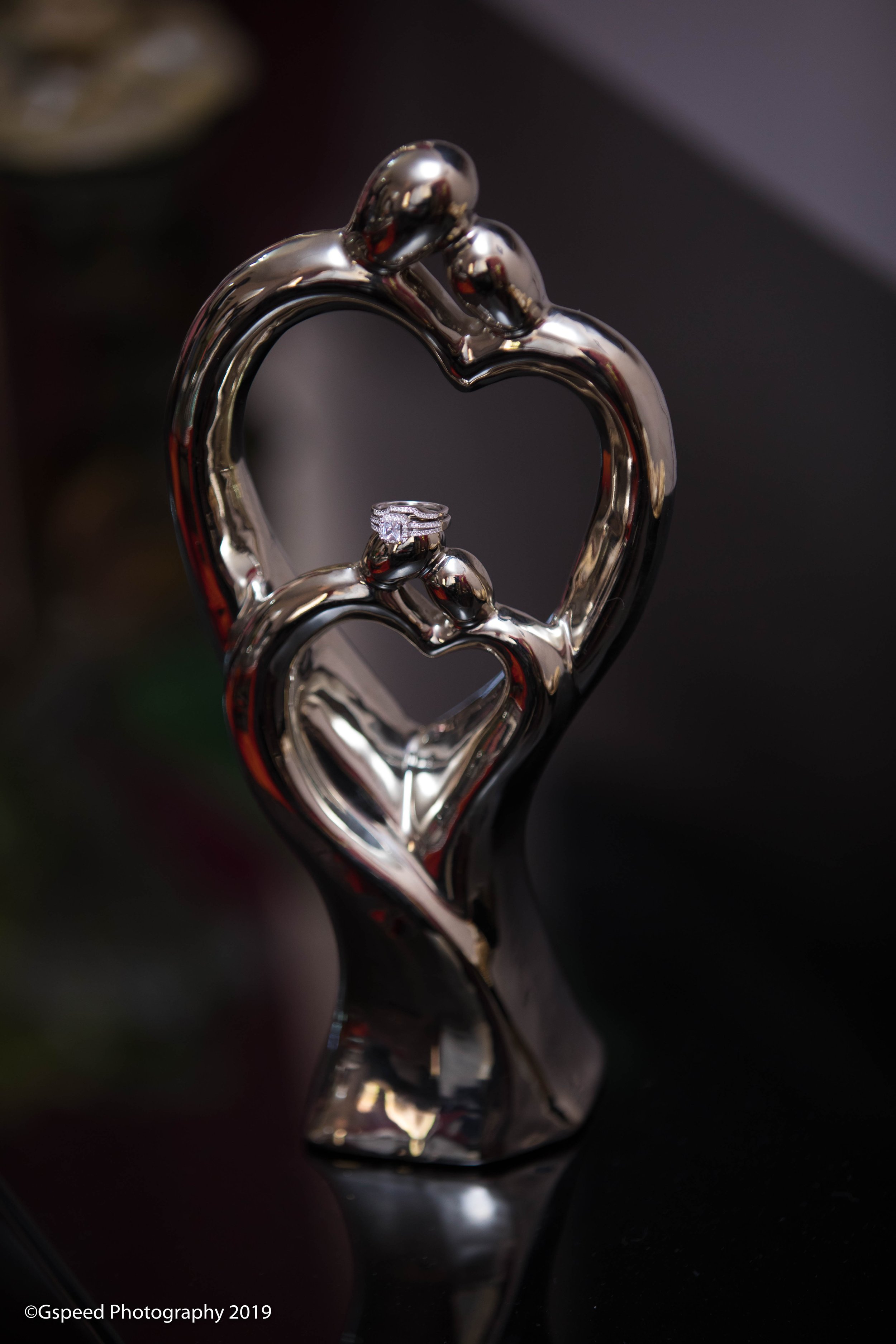 A silver heart-shaped metal sculpture with two small ball figures on top, resembling hugging or leaning towards each other, set against a dark blurred background.