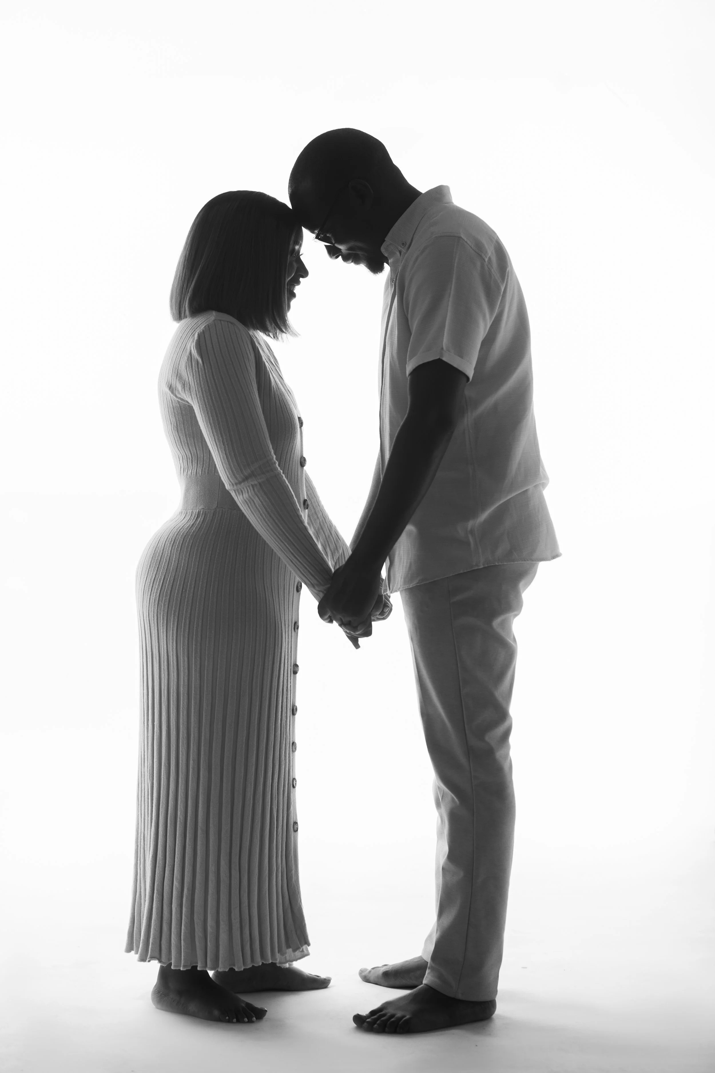creative studio photoshoot of A black-and-white photo of a couple holding hands, with their foreheads touching, looking affectionately at each other, standing barefoot against a plain white background.